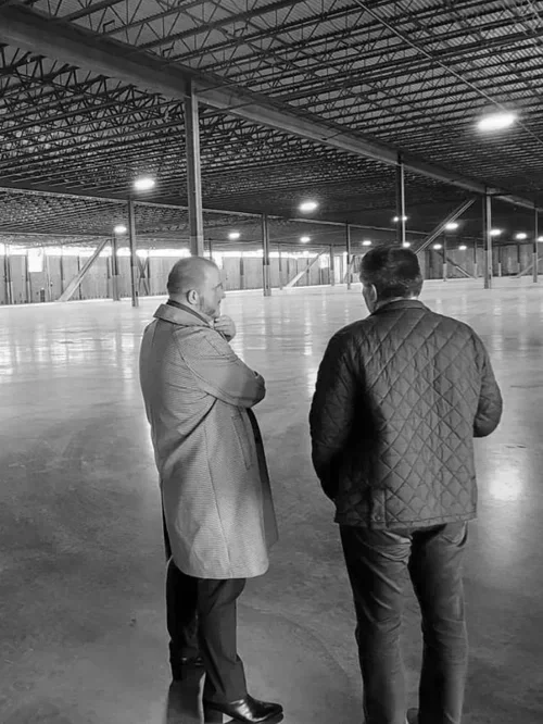 Two men standing inside a large, empty warehouse with a high ceiling and steel support beams, engaged in conversation.