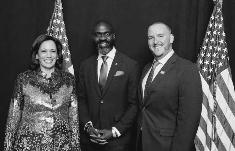 Three individuals in formal attire standing in front of U.S. flags, smiling for a photo.