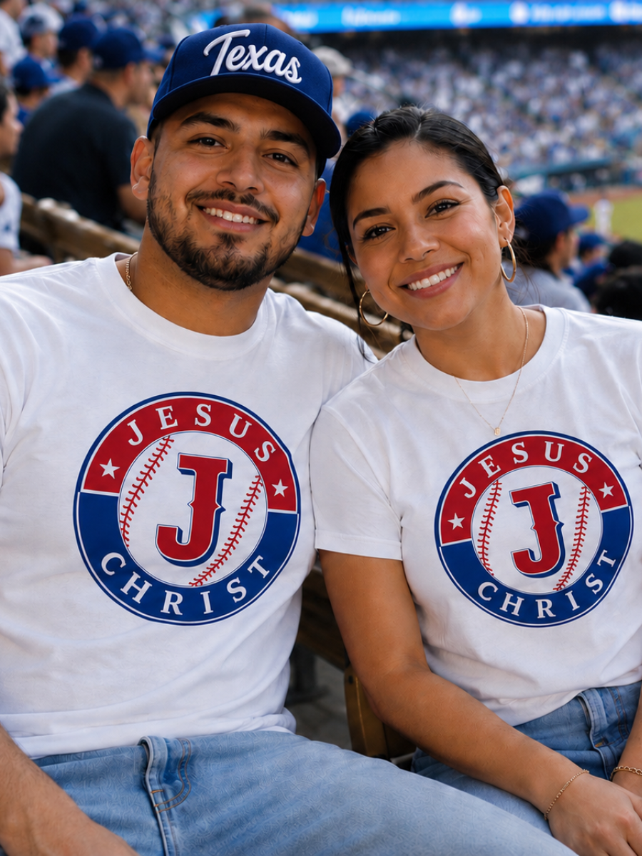 A smiling man and woman sit in stadium bleachers, wearing white t-shirts with a "Jesus Christ" logo featuring a baseball and a large J, at a baseball game.