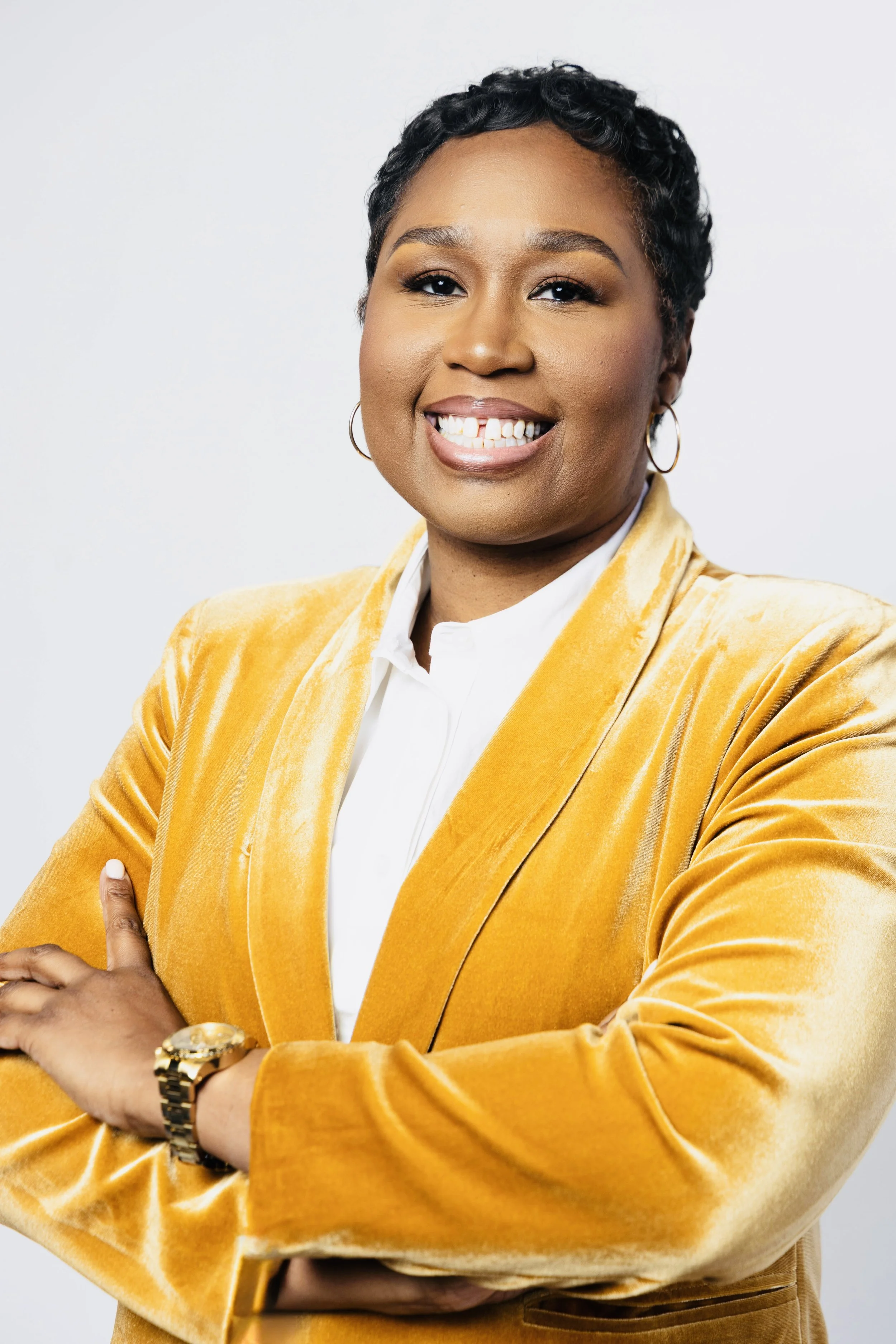 A woman with short curly black hair wearing a yellow velvet blazer, white shirt, and gold jewelry, smiling with arms crossed against a plain white background.
