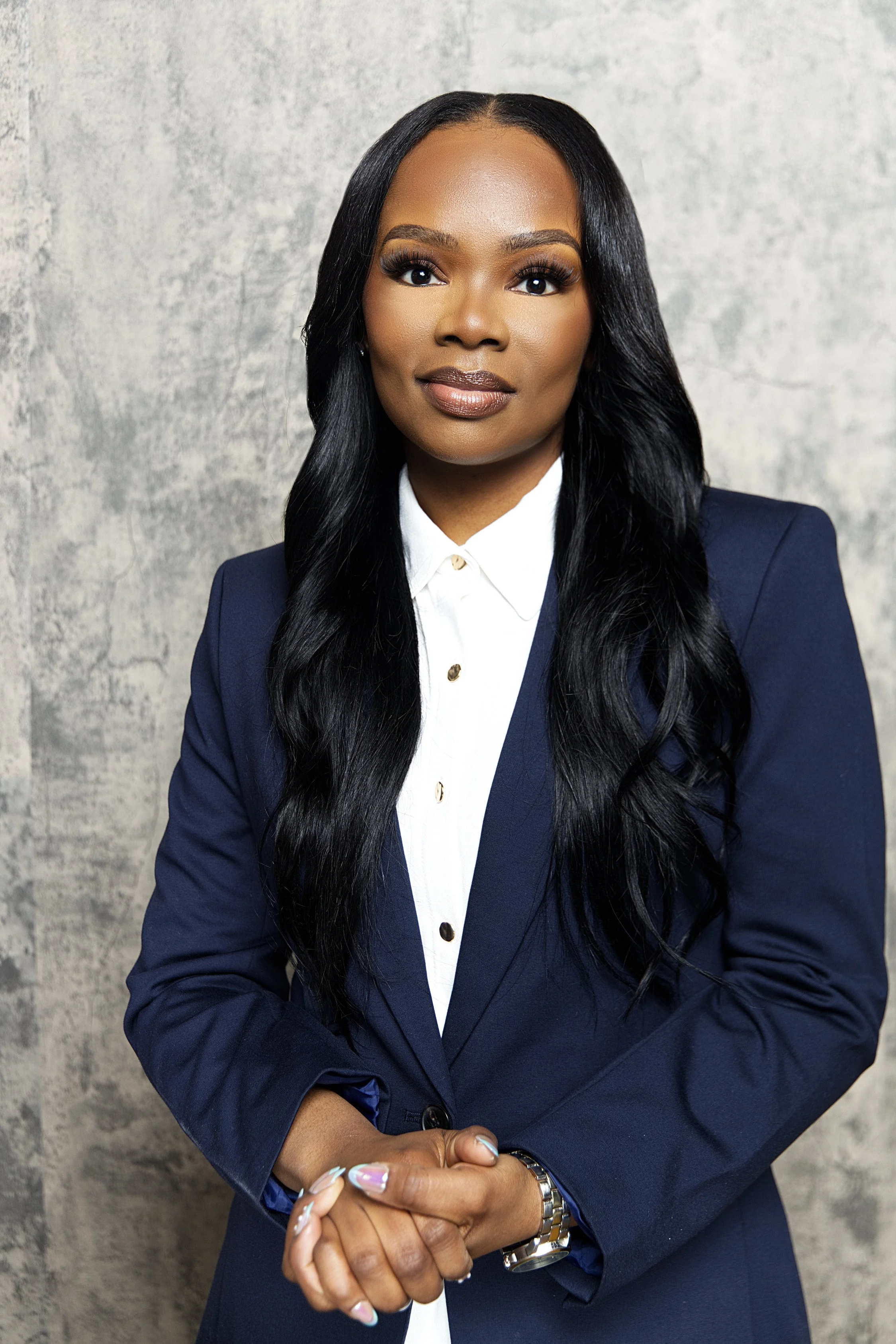 Portrait of a Black woman in a navy blazer and white shirt, standing against a gray textured background.