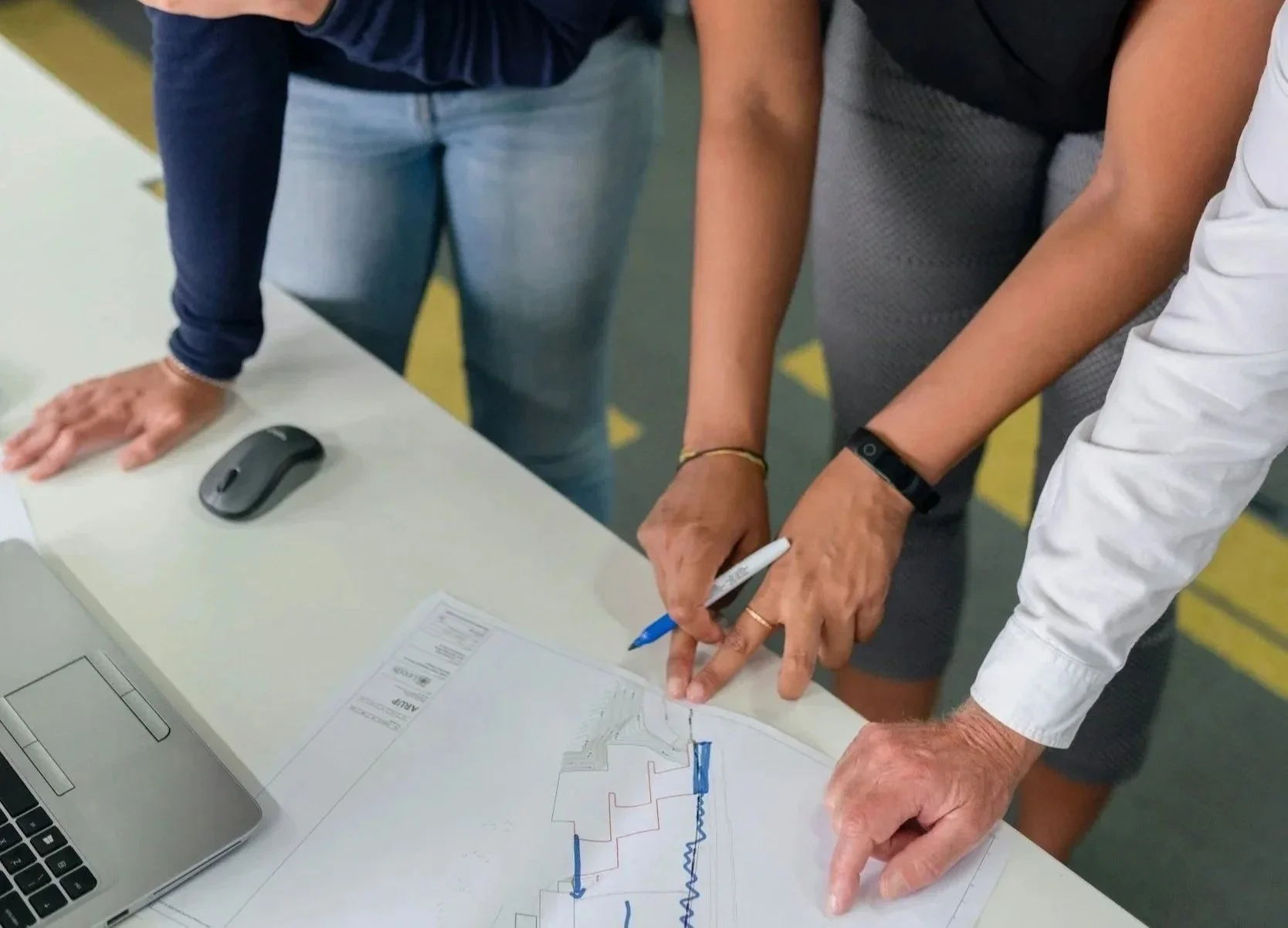 Two people are collaborating over a map or diagram on a table, pointing at it with a pen. One person's hand is on the table, another's is holding a pen and pointing to the map. A laptop and a computer mouse are on the table.