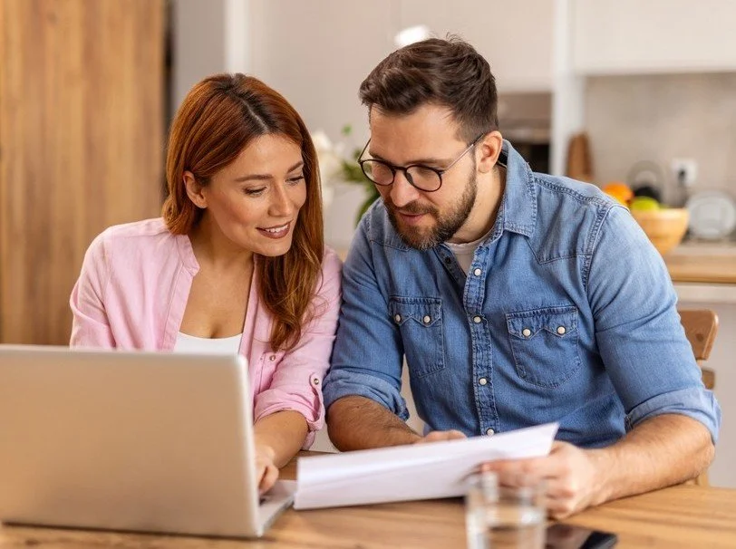 A woman and man reviewing documents together in a kitchen, with a laptop on the table.