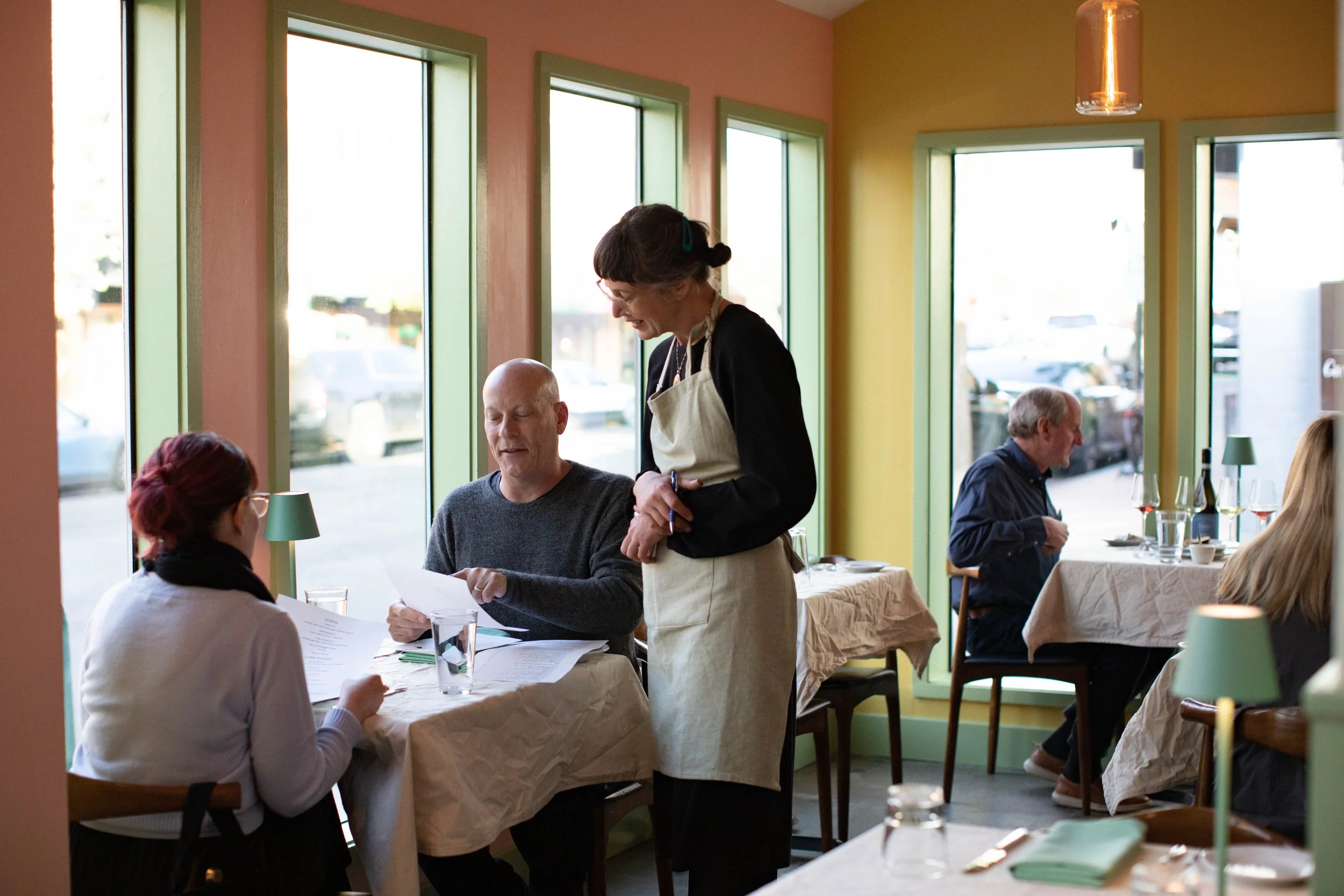 a server providing warm, welcoming hospitality to guests who are excited to dine. soft colors make the walls and a warm glow lights the restaurant. There are tablecloths and polished silverware as well as bottles of wine on the tables.