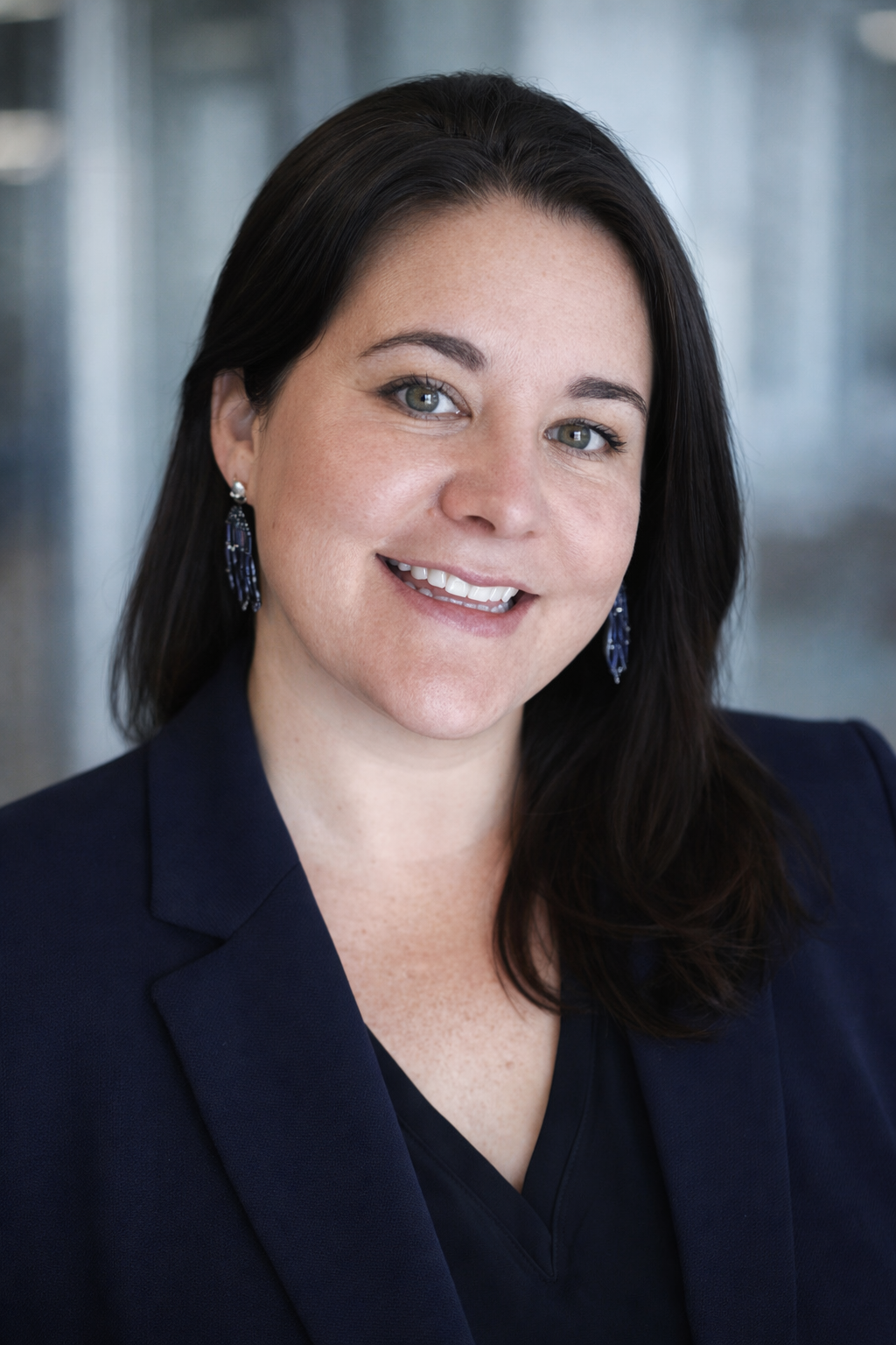 Professional headshot of a woman with dark hair, blue eyes, wearing a navy blazer and blue earrings, smiling in front of a blurred background.