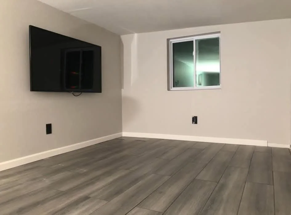 Empty room with gray wood floors, light-colored walls, a mounted flat-screen TV, a window, and electrical outlets.