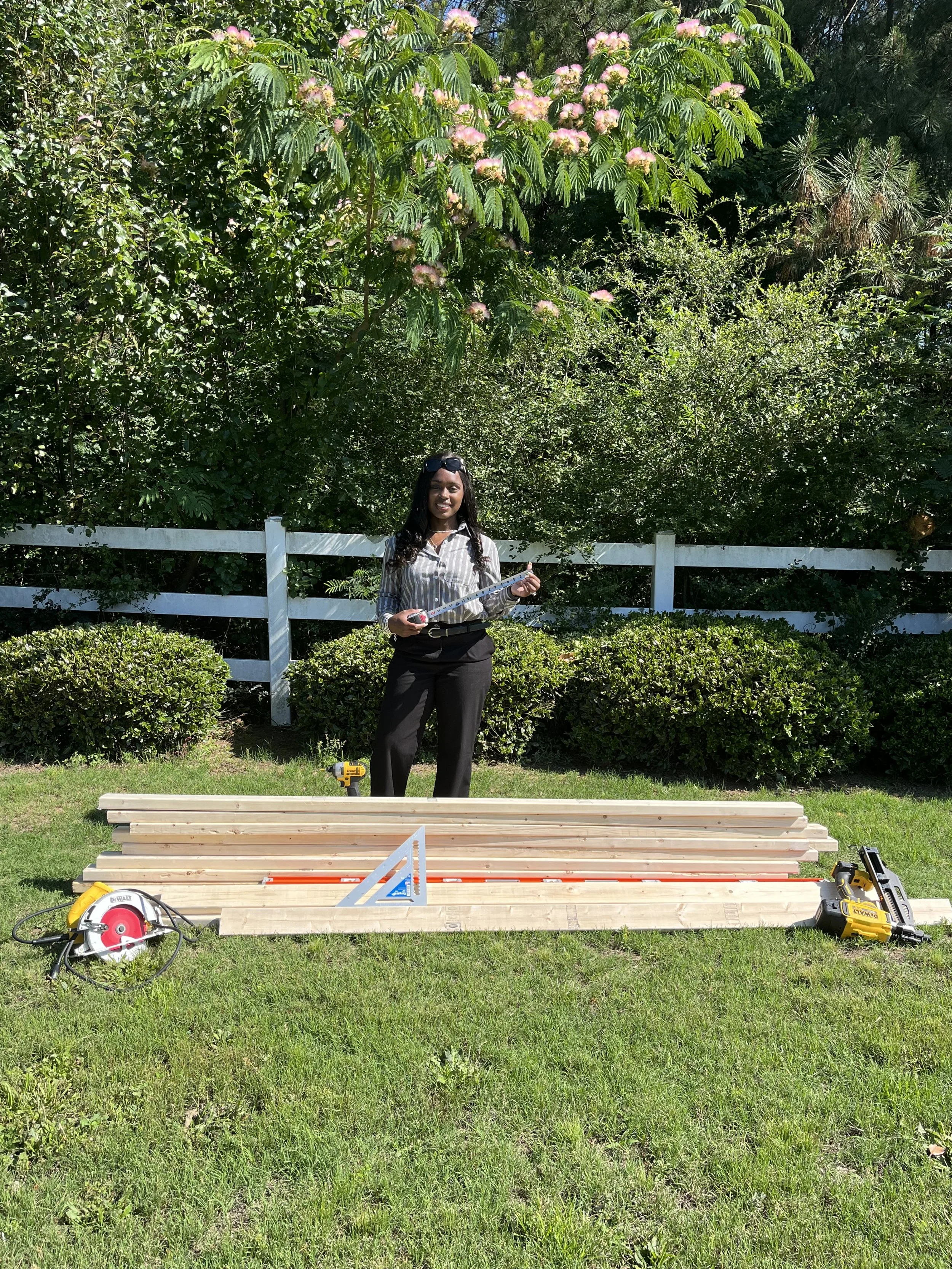 A woman holding a measuring tape smiling outdoors in front of a construction project with wooden planks, power tools, and a saw on the grass. There is a white fence and lush greenery with pink flowering trees in the background.