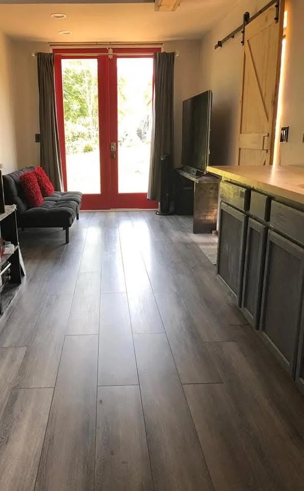 Living room with a black sofa, a TV on a stand, and large glass doors with red trim leading outside, wooden sliding barn door on the wall, and gray flooring.