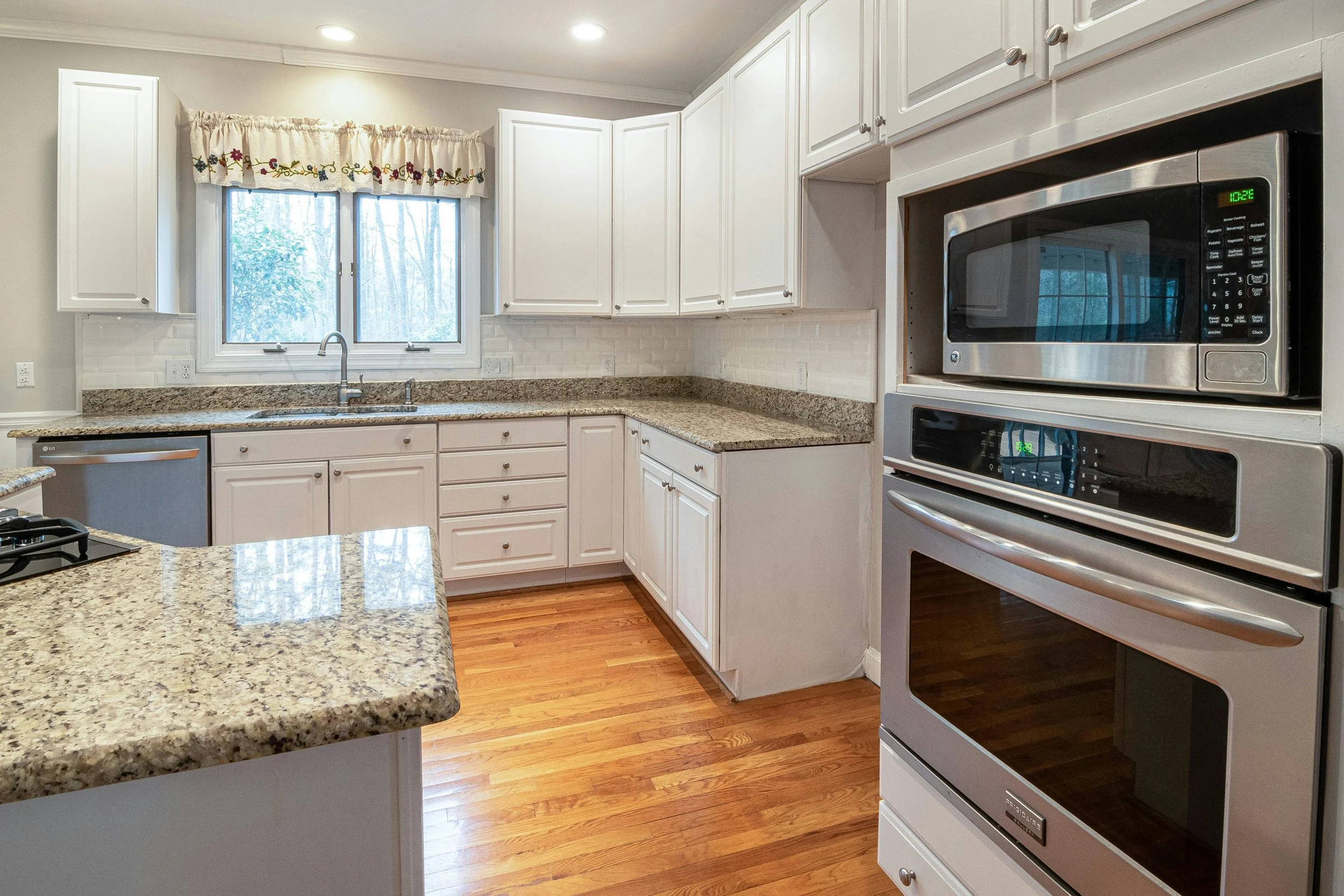 A modern kitchen with white cabinets, granite countertops, stainless steel microwave, and oven, hardwood flooring, a window with floral curtains, and a faucet over the sink.