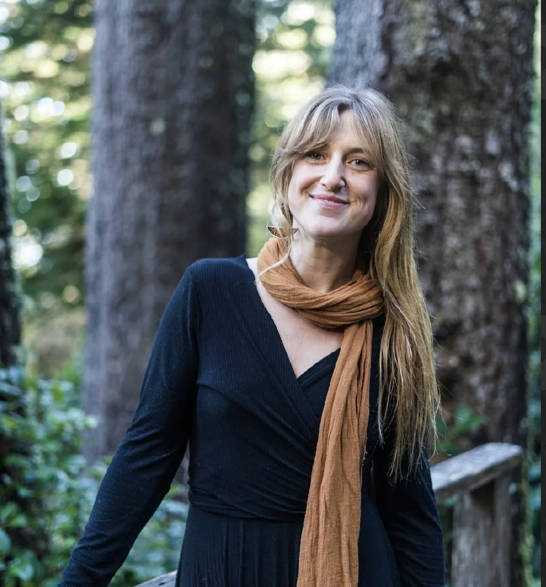 A smiling woman in a black dress and brown scarf standing outdoors in front of large trees.