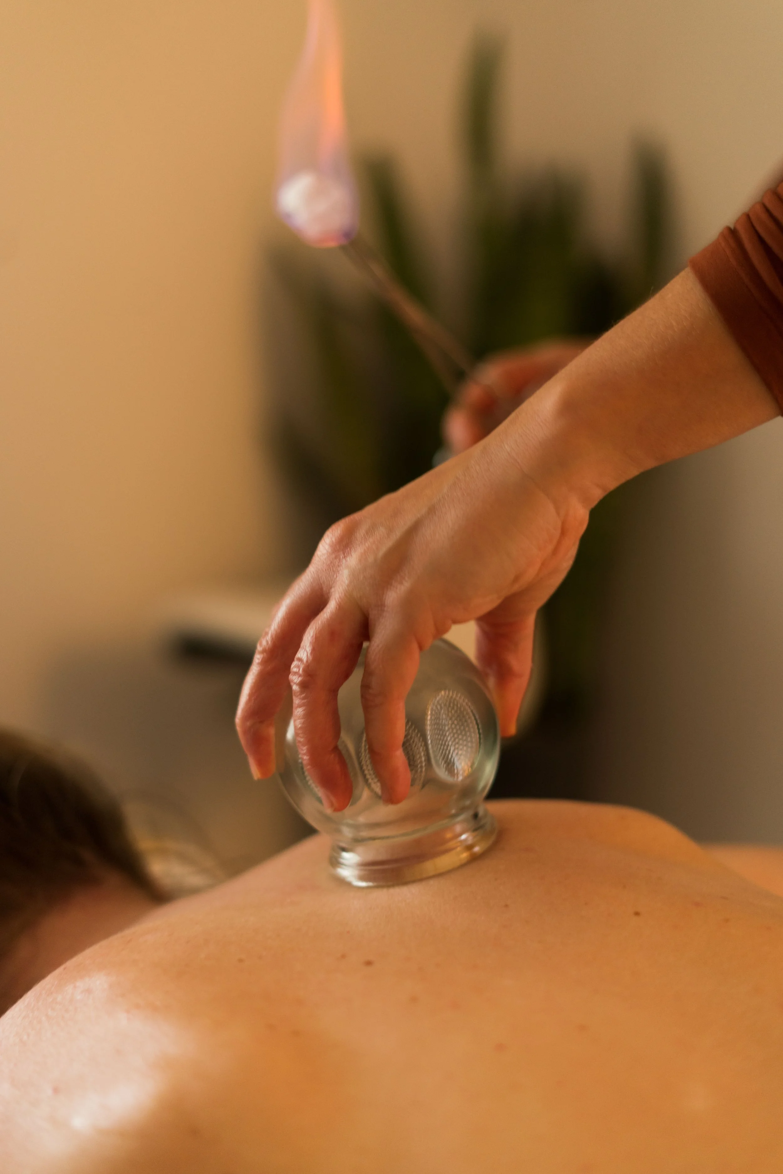 Close-up of a person receiving a massage or therapy with a glass cup on their back and a practitioner’s hand holding a small metal object, possibly for cupping or acupuncture, with blurred background.