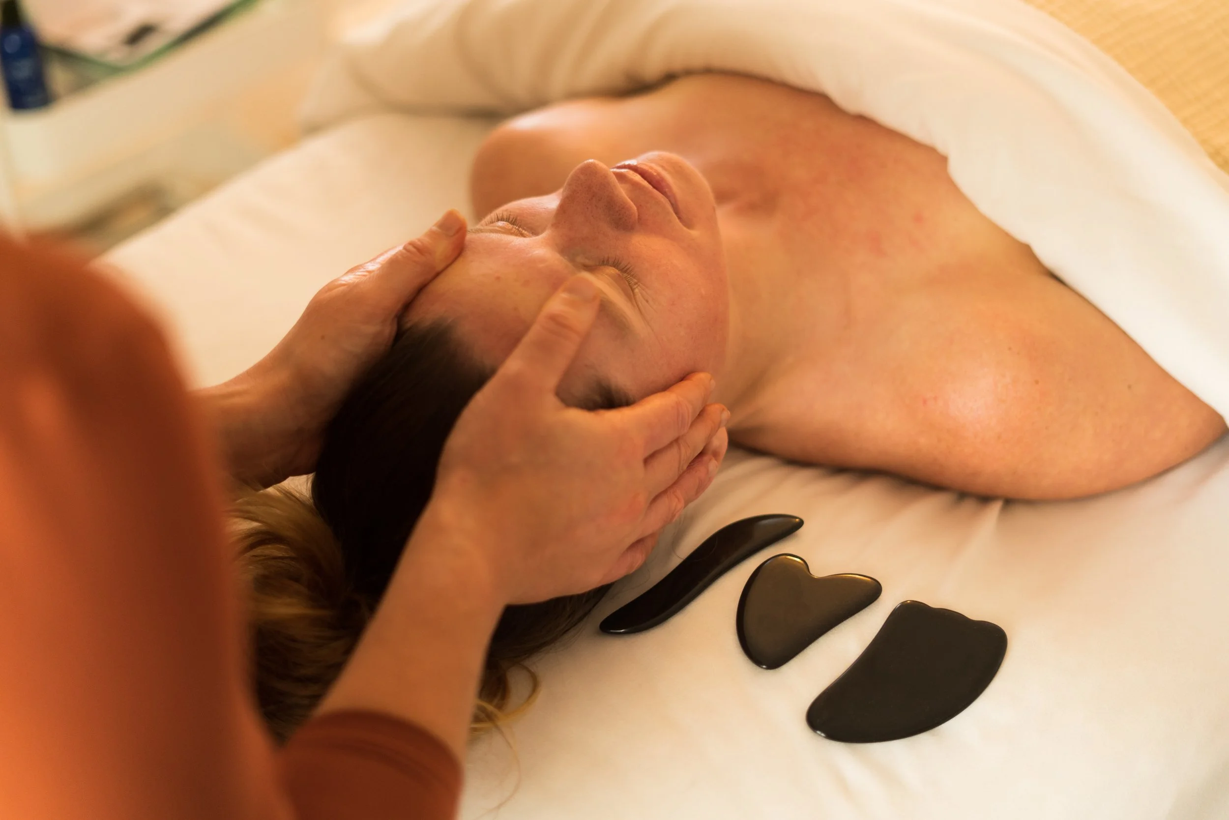 Person receiving a facial massage in a spa or skincare setting, with black facial stones arranged on a white towel nearby.