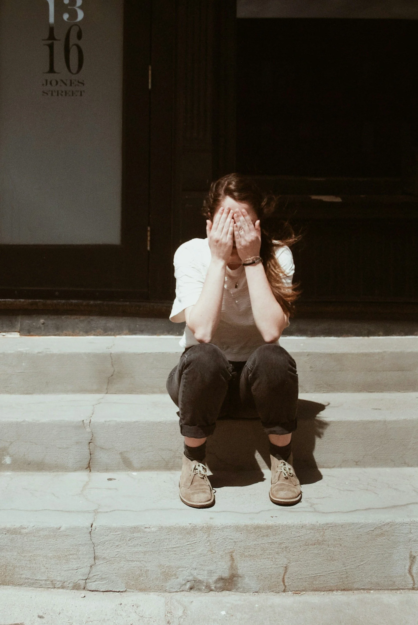 A woman sitting on concrete steps covering her face with her hands, wearing a white shirt, black pants, beige shoes, and black socks, after sunset with a dark background and a street sign reading '13 16 JONES STREET' on the wall to the left.