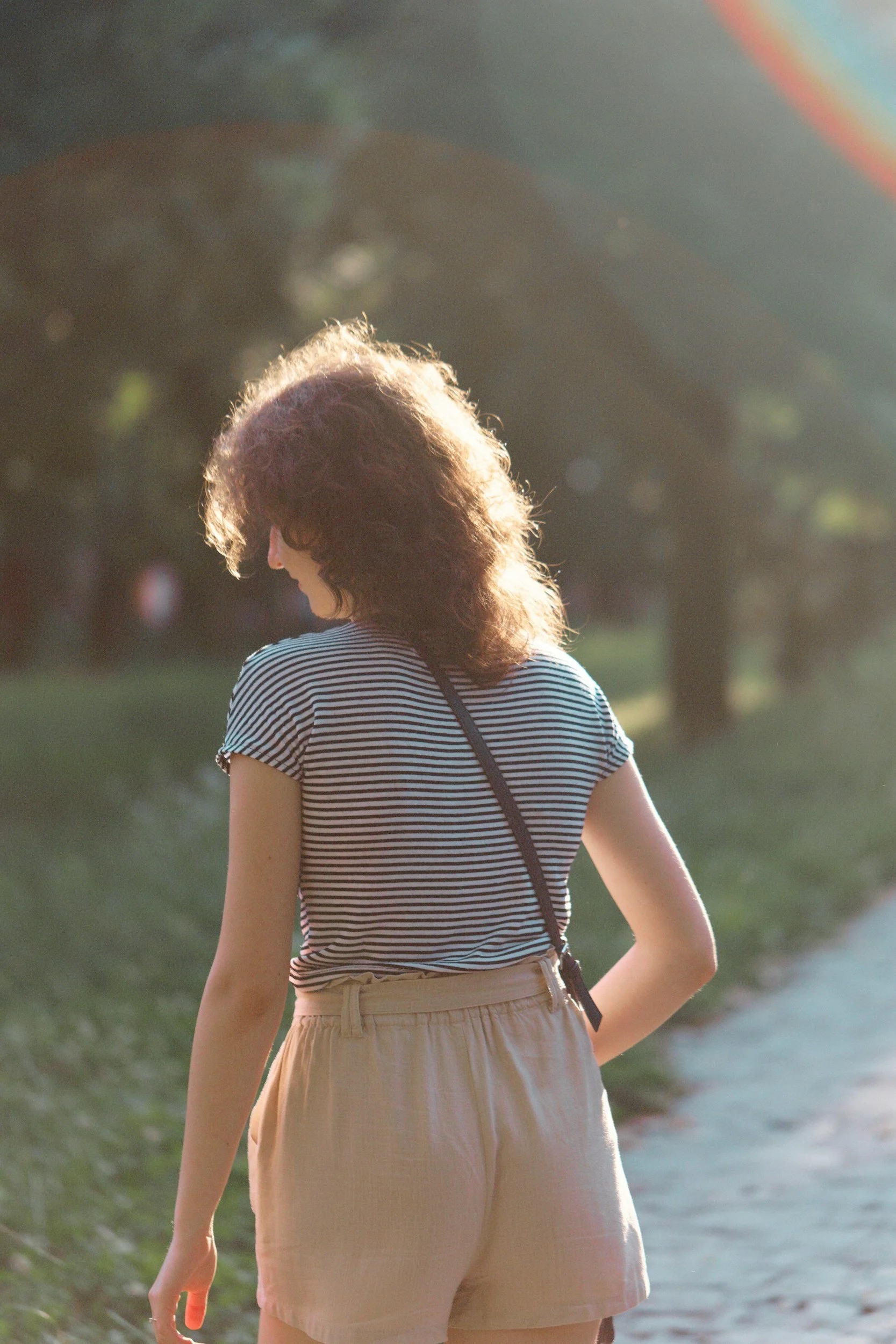 A woman with curly hair wearing a striped T-shirt and beige shorts standing outdoors near water, back turned to the camera, with trees and sunlight in the background.
