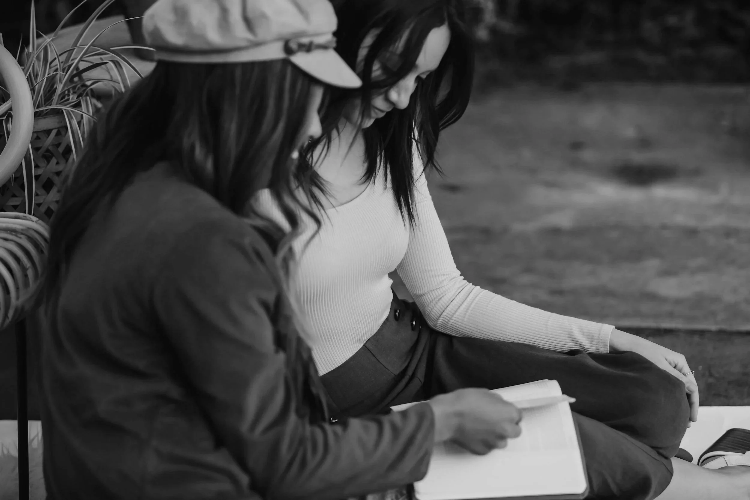 A black and white photo of two women sitting next to each other, engaged in reading or writing. One woman has long dark hair, glasses, a light-colored top, and dark pants. The other woman’s face is not visible, but she is wearing a jacket. They are seated on a bench or similar surface, with some plants visible behind them.