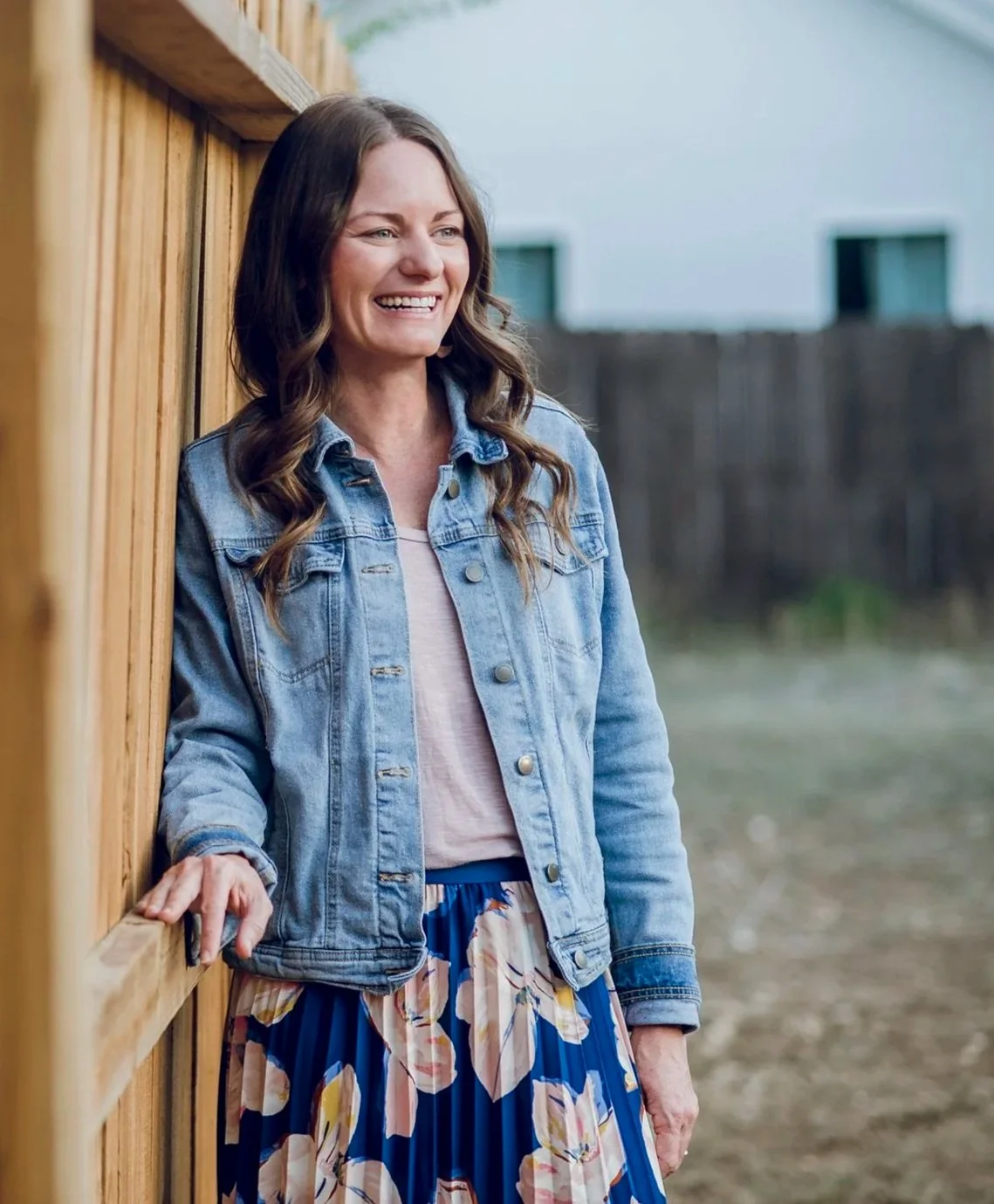 A woman, Lee Anne Bingham, with brown hair styled in loose waves, smiling, standing outdoors leaning against a wooden fence. She is wearing a light pink top, a denim jacket, and a blue floral skirt.