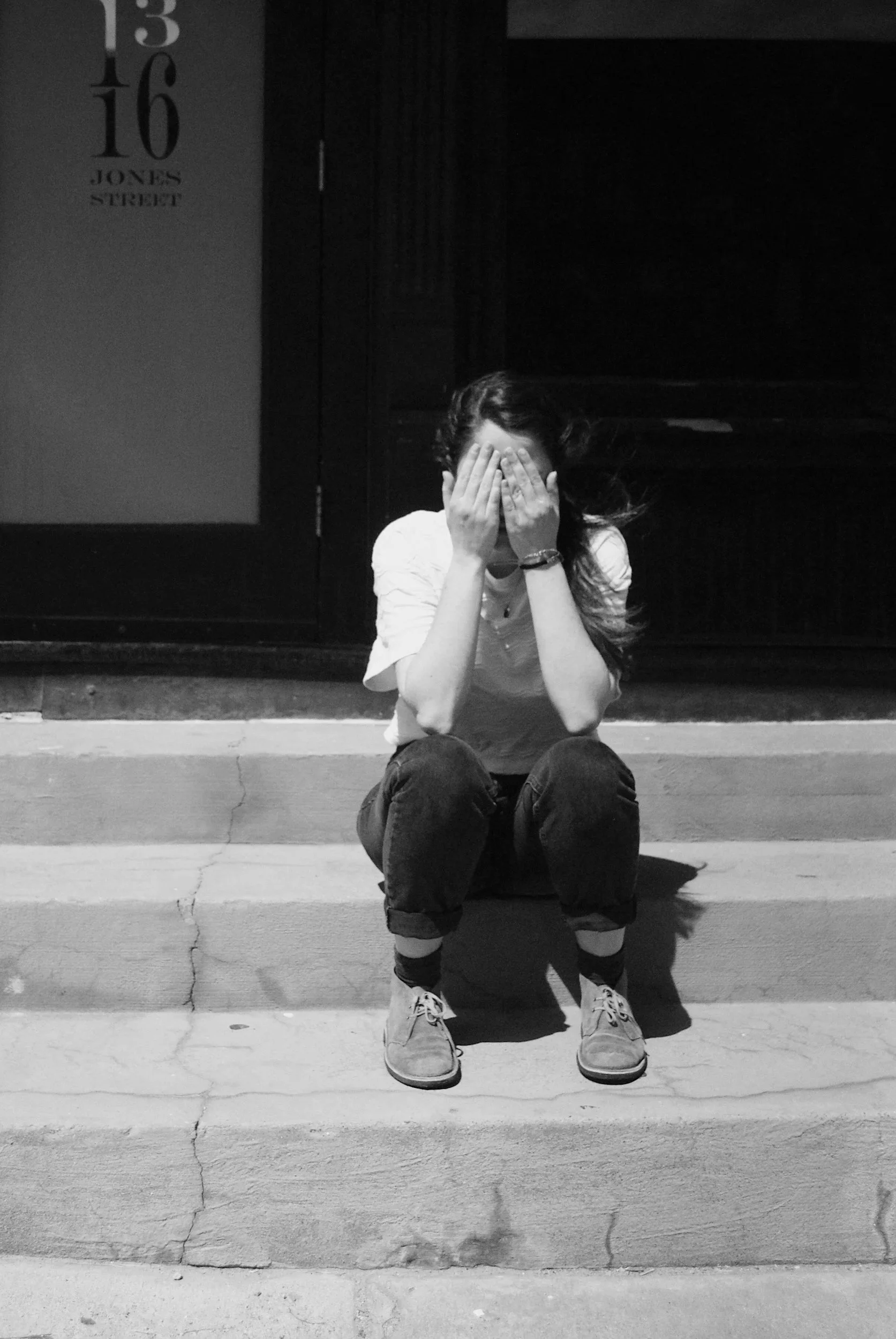Black and white photo of a person sitting on outdoor stairs with their legs apart and their hands covering their face. The person is wearing a light-colored t-shirt, dark pants, and sneakers. There is a sign on the wall behind them with the address "13 16 Jones Street.