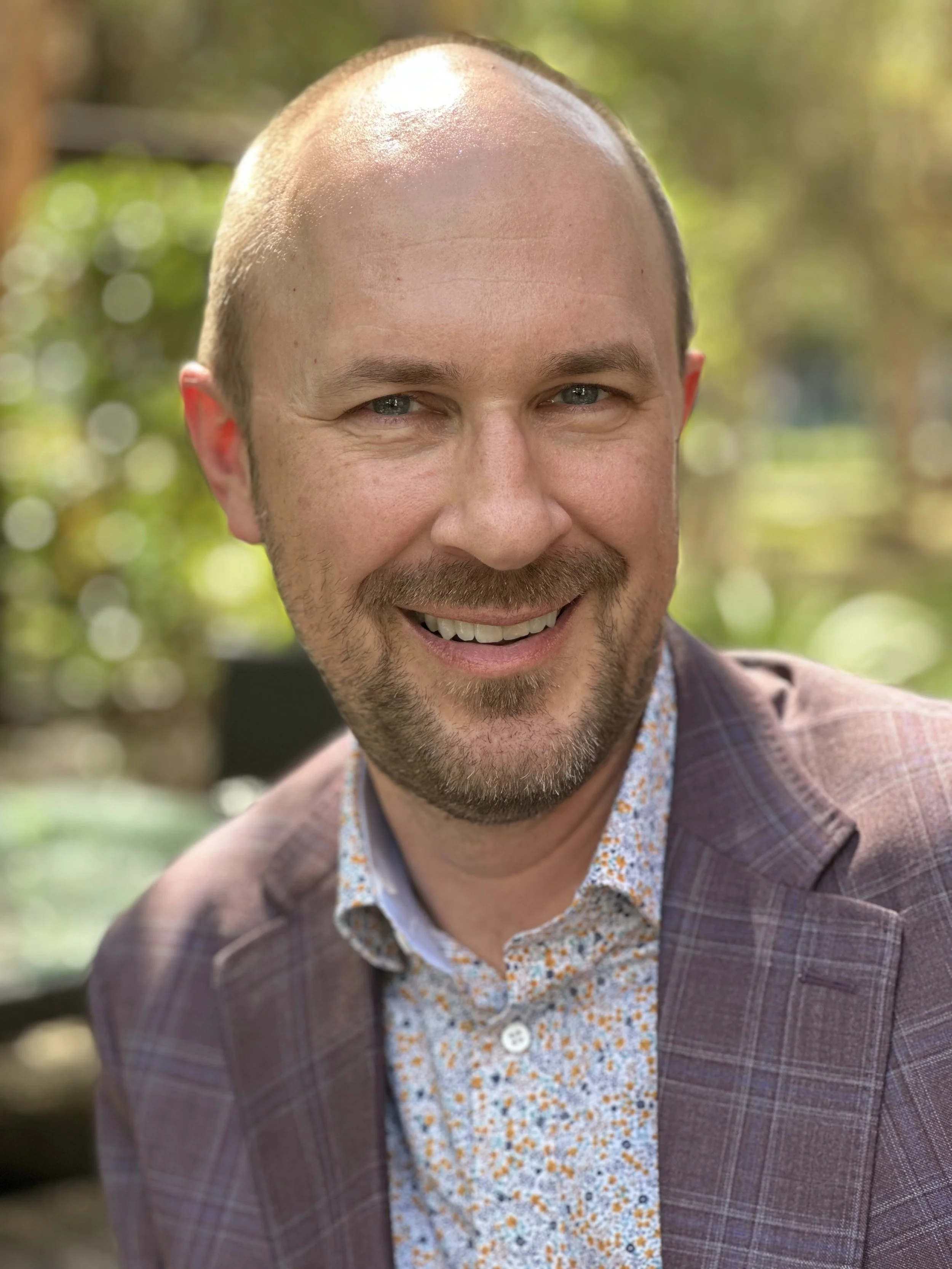 Close-up of a smiling man with a beard, wearing a brown plaid blazer and a floral shirt, outdoors with blurred green background.