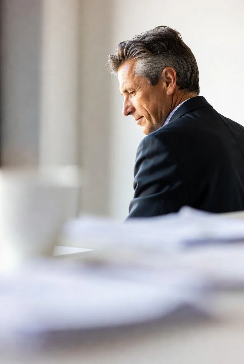 A middle-aged man in a dark suit is sitting at a desk with papers in front of him, looking pensively out a window.
