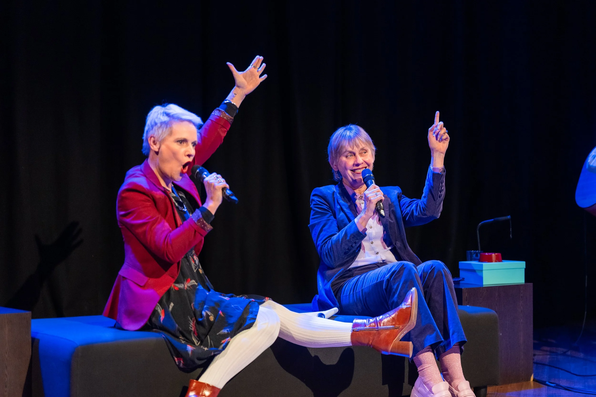 Two women speaking into microphones on stage, one with short gray hair and a red blazer, the other with short light brown hair and a navy blazer, sitting on a bench with colorful objects beside them.