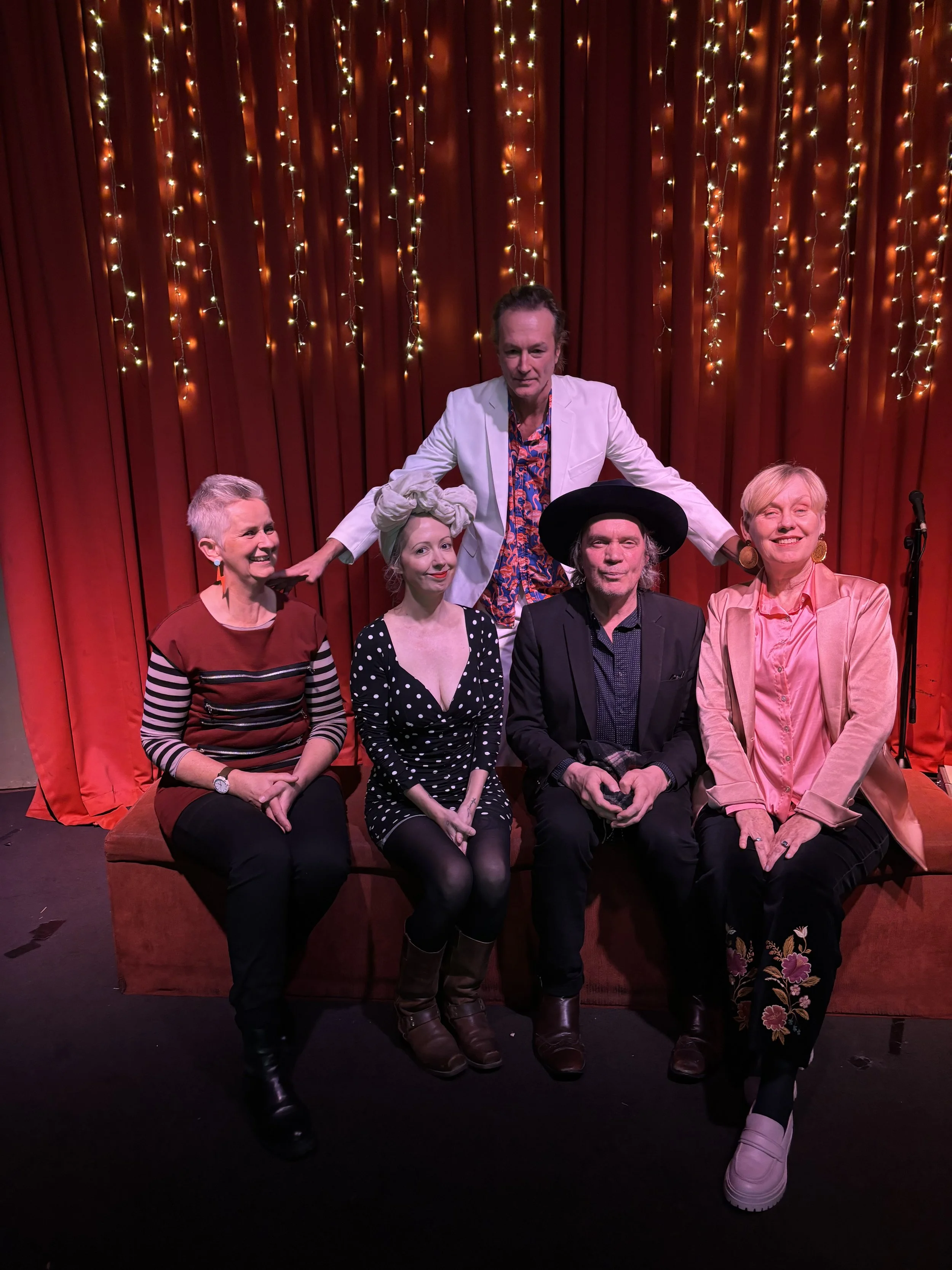Five people posing in front of red curtains with string lights. One man stands behind the others, wearing a white blazer and colorful shirt. The four women and one man sitting in front are dressed in stylish clothing, with one woman wearing a head wr