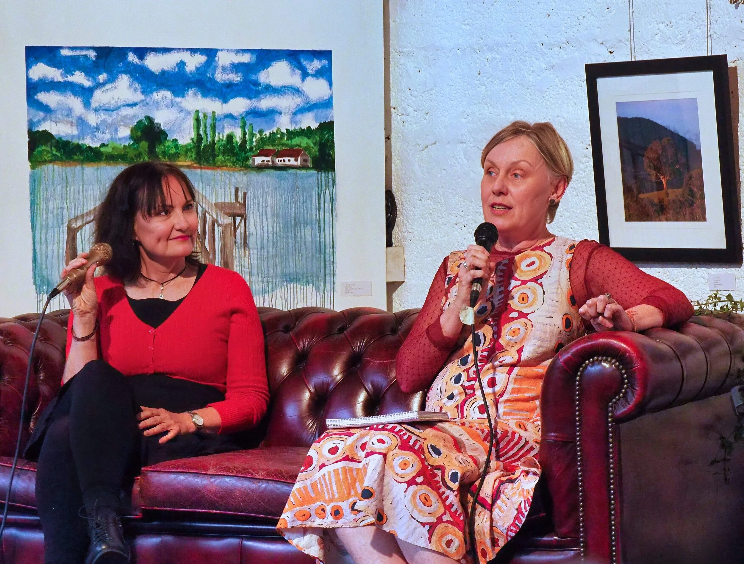 Two women sitting on a burgundy leather sofa and holding microphones at an art gallery, with artwork hanging on the walls behind them.