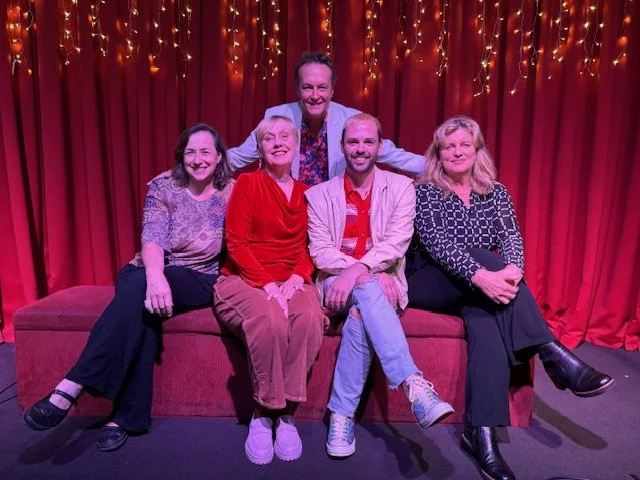 Group of five people sitting on a red couch in front of a red curtain with string lights, smiling for the camera, in a stage setting.