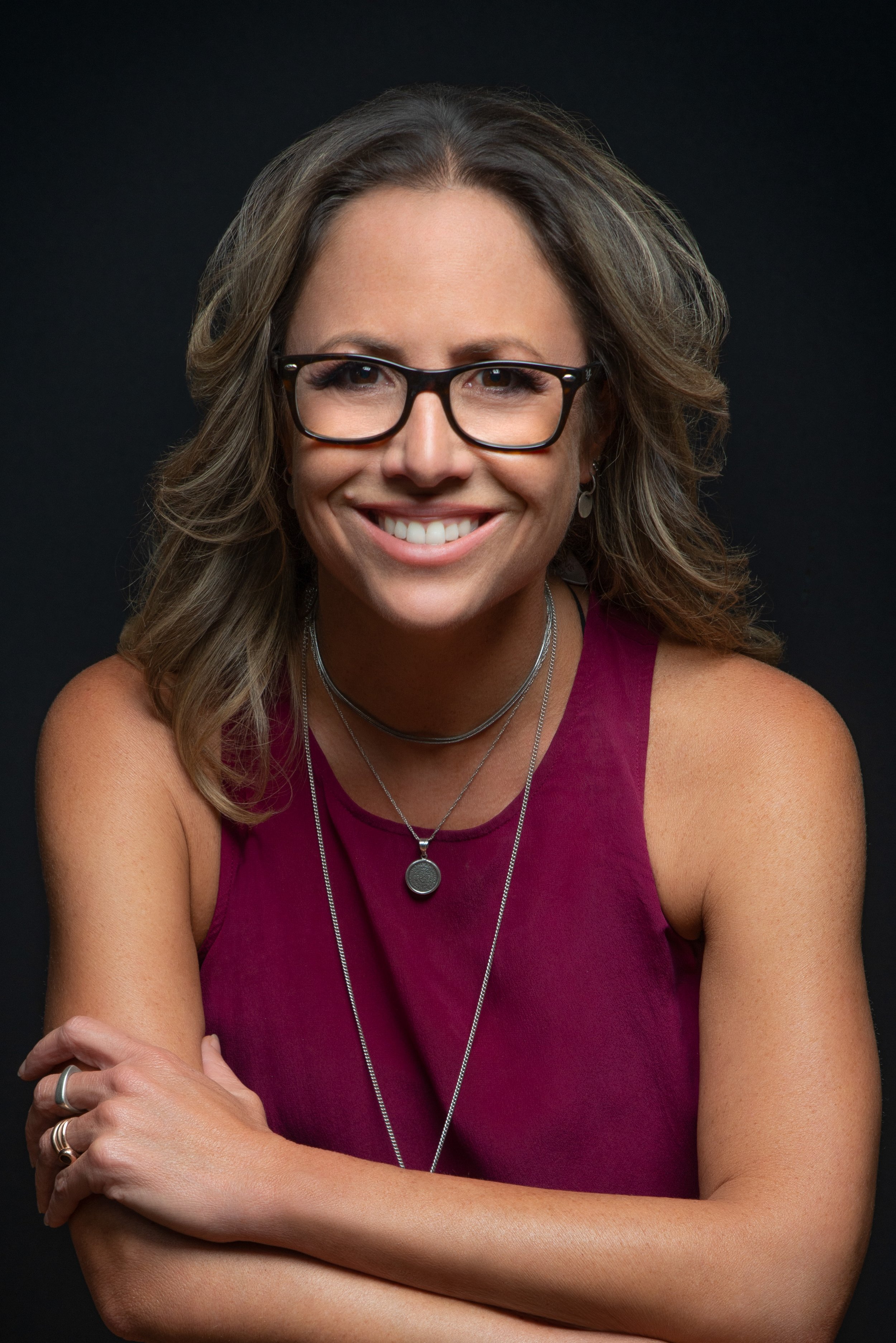 A woman with shoulder-length wavy hair, wearing glasses, a sleeveless burgundy top, and layered necklaces, smiling at the camera against a black background.