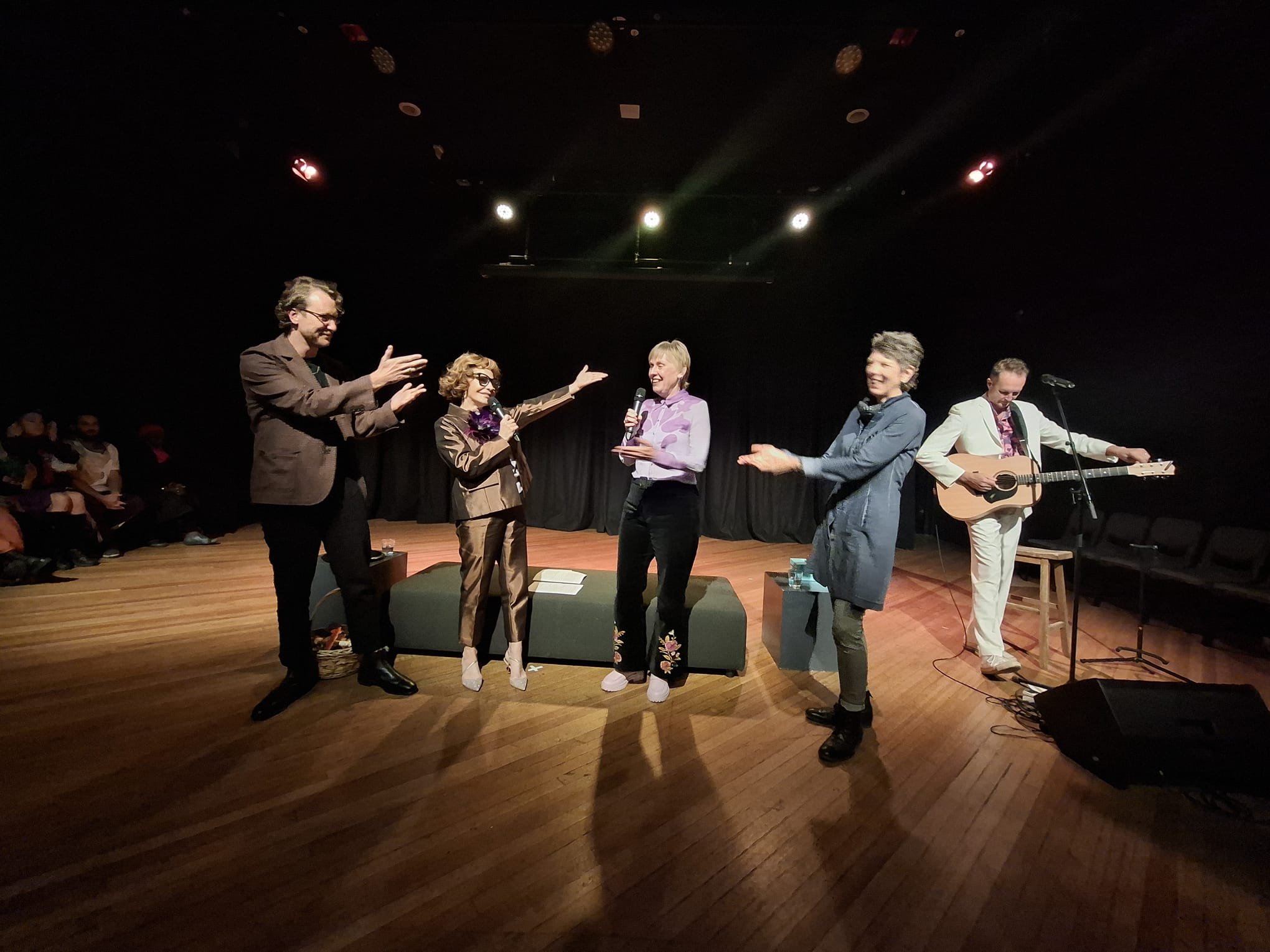 Group of five people on stage, some holding microphones, one with guitar, engaged in a performance or presentation, with an audience watching.