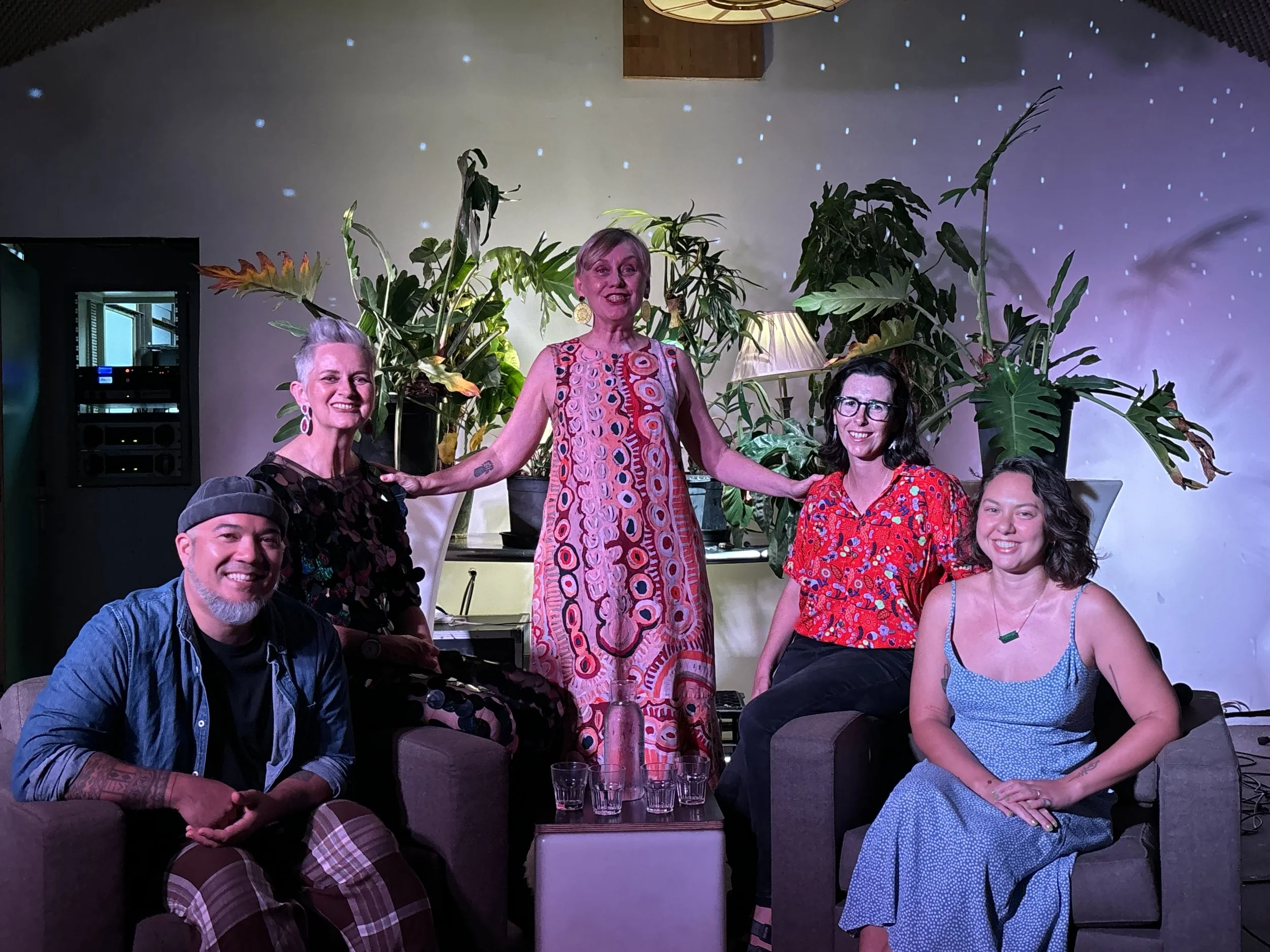 Group of seven people, five women and two men, posing together indoors with a backdrop of large green plants and a starry night projection on the wall.