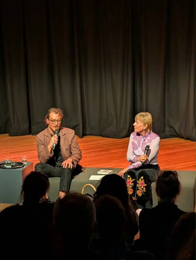 A man and a woman are seated on a stage in front of an audience, participating in a discussion or interview. The man holds a microphone, wears glasses and a blazer, and the woman also holds a microphone, wears a light-colored blouse with purple patte