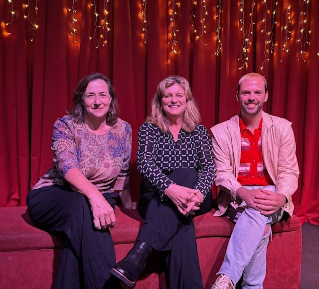 Three people sitting on a red stage with a red curtain and string lights behind them.
