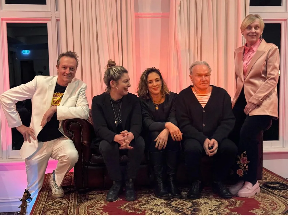 Group of five people posing on a sofa in a warmly lit room, with pink curtains and a patterned rug.