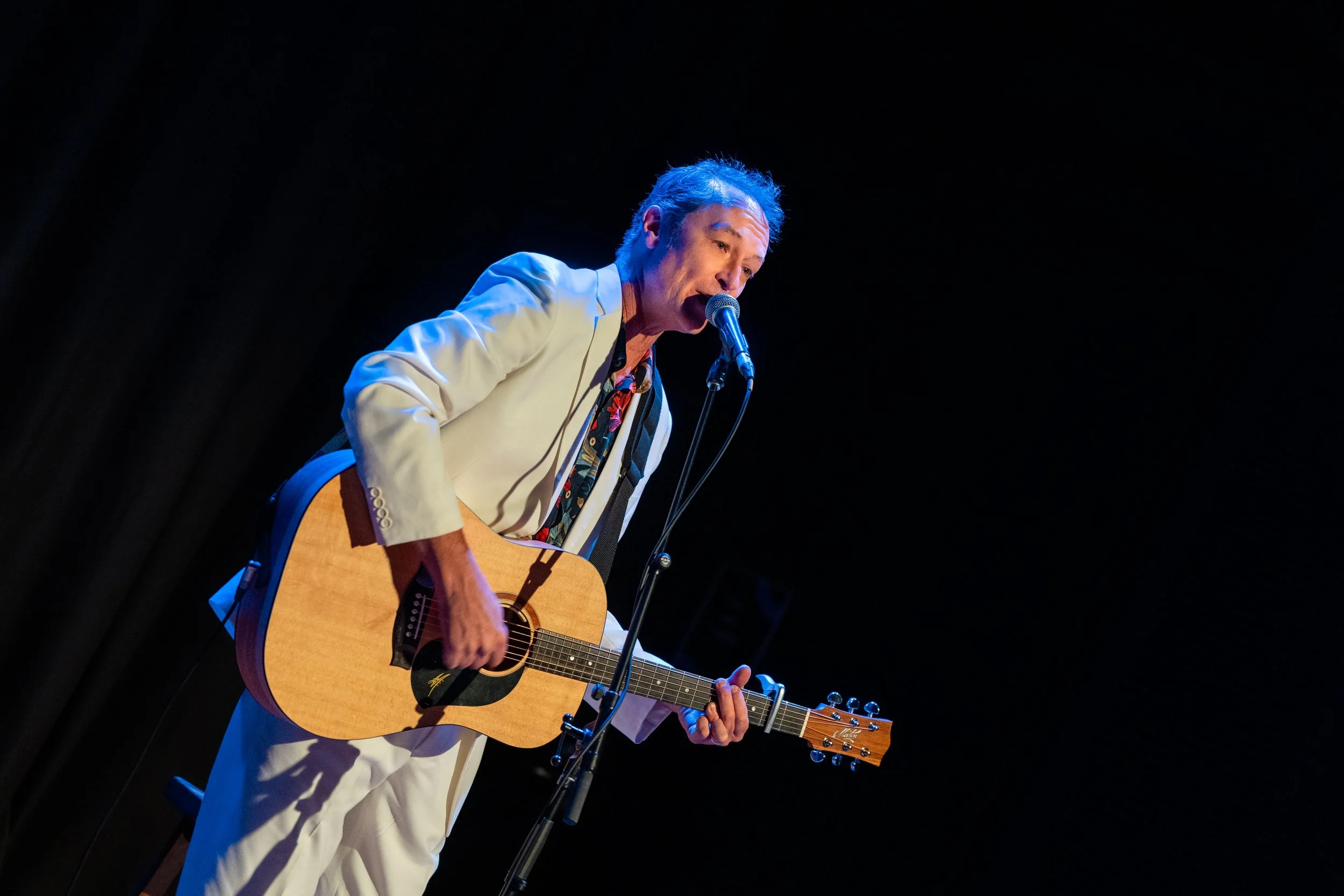A man with gray hair wearing a white suit and patterned shirt is playing an acoustic guitar and singing into a microphone on a dark stage.
