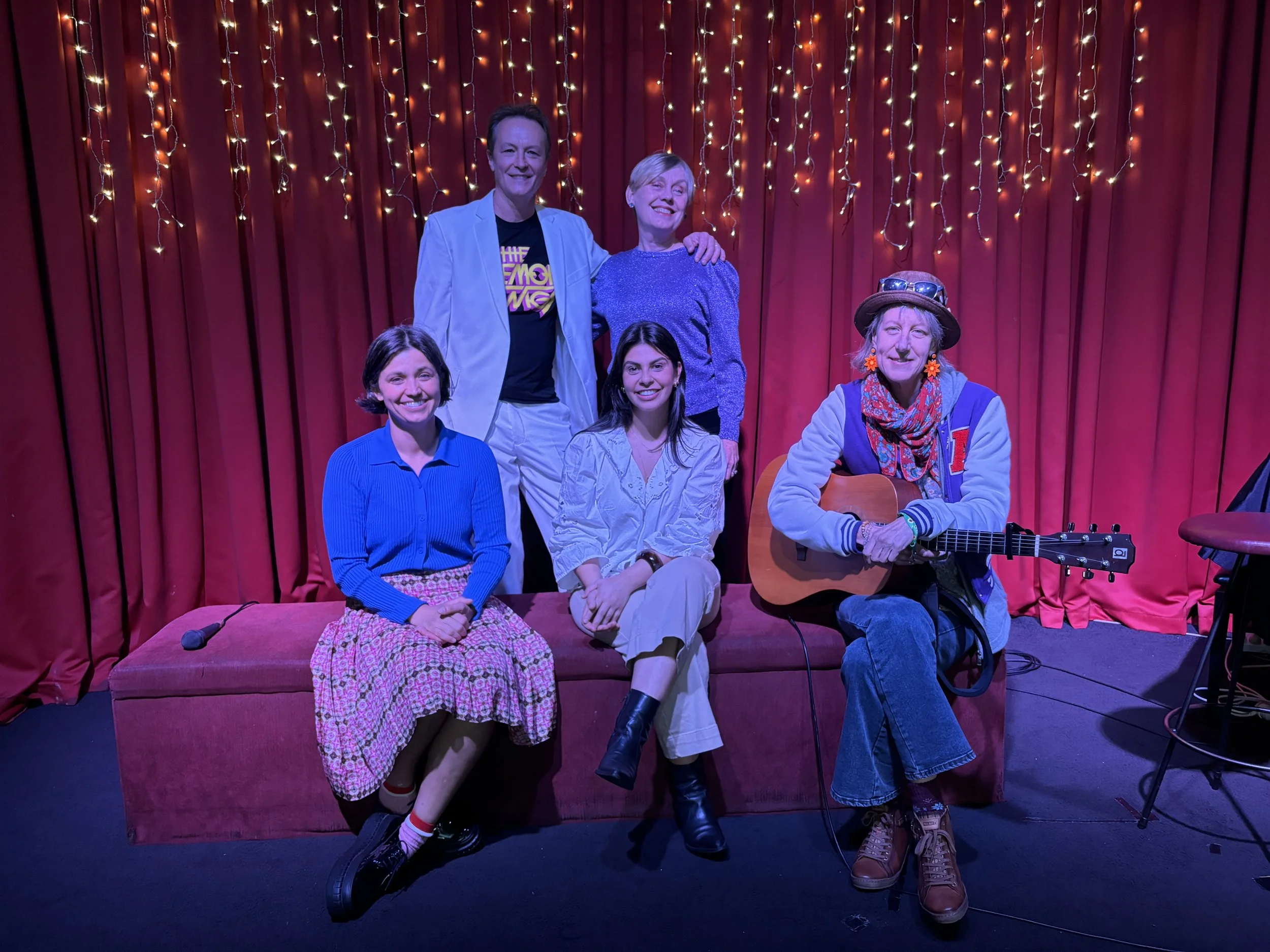 Group of five smiling people on stage with red curtains and string lights in the background. They are seated and standing, some with musical instruments and a microphone.