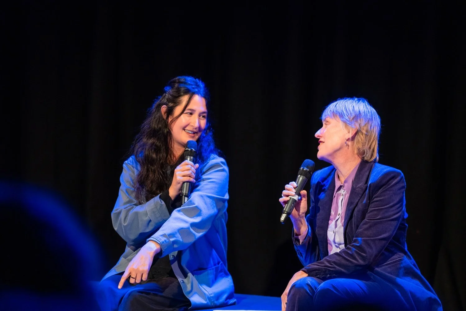 Two women sitting on stage, holding microphones and engaging in conversation, with a black curtain backdrop.