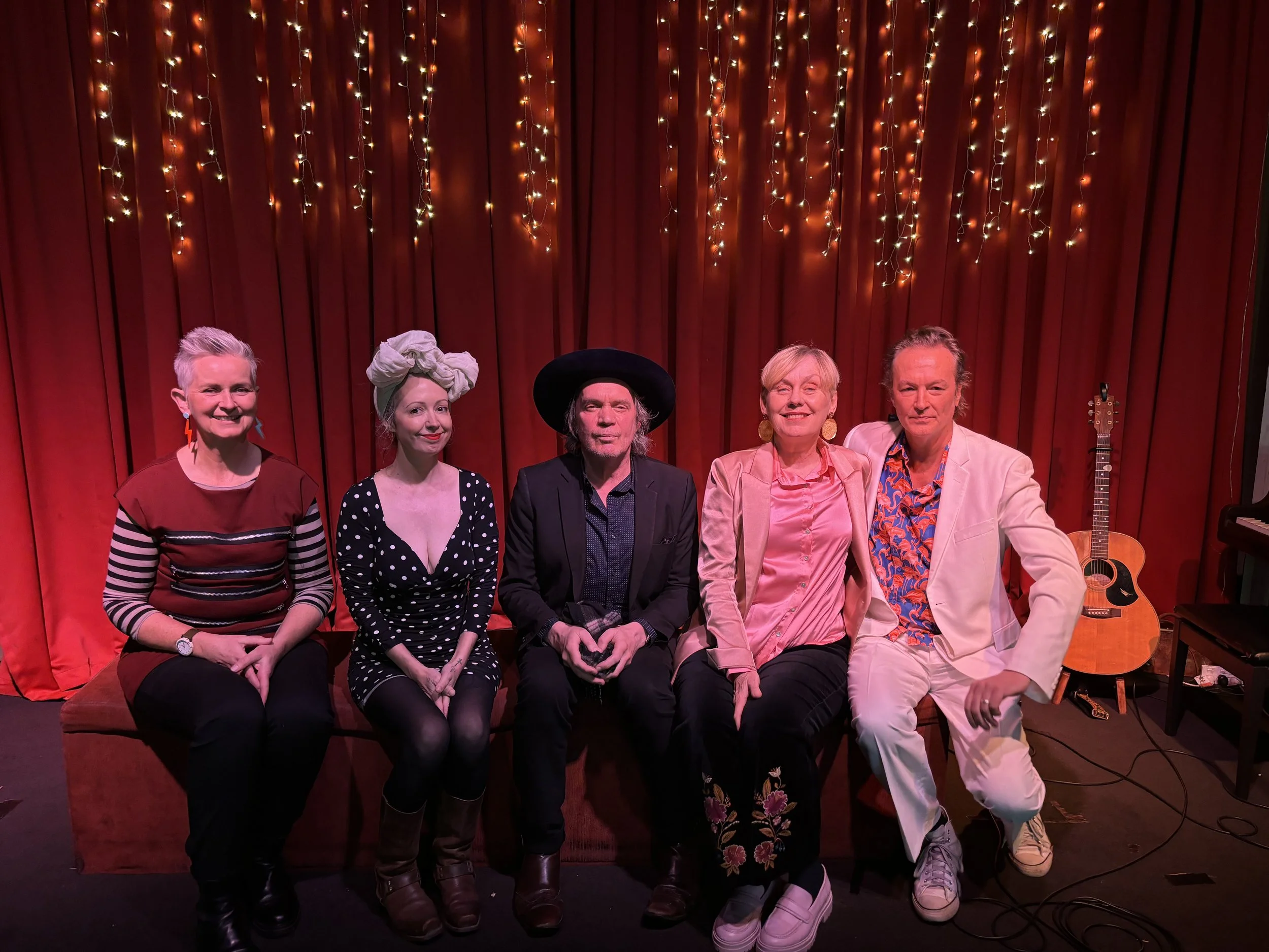 Five people sitting on a bench in front of a red curtain with hanging string lights. They are smiling and dressed in colorful clothing, with a guitar visible on the right side of the image.
