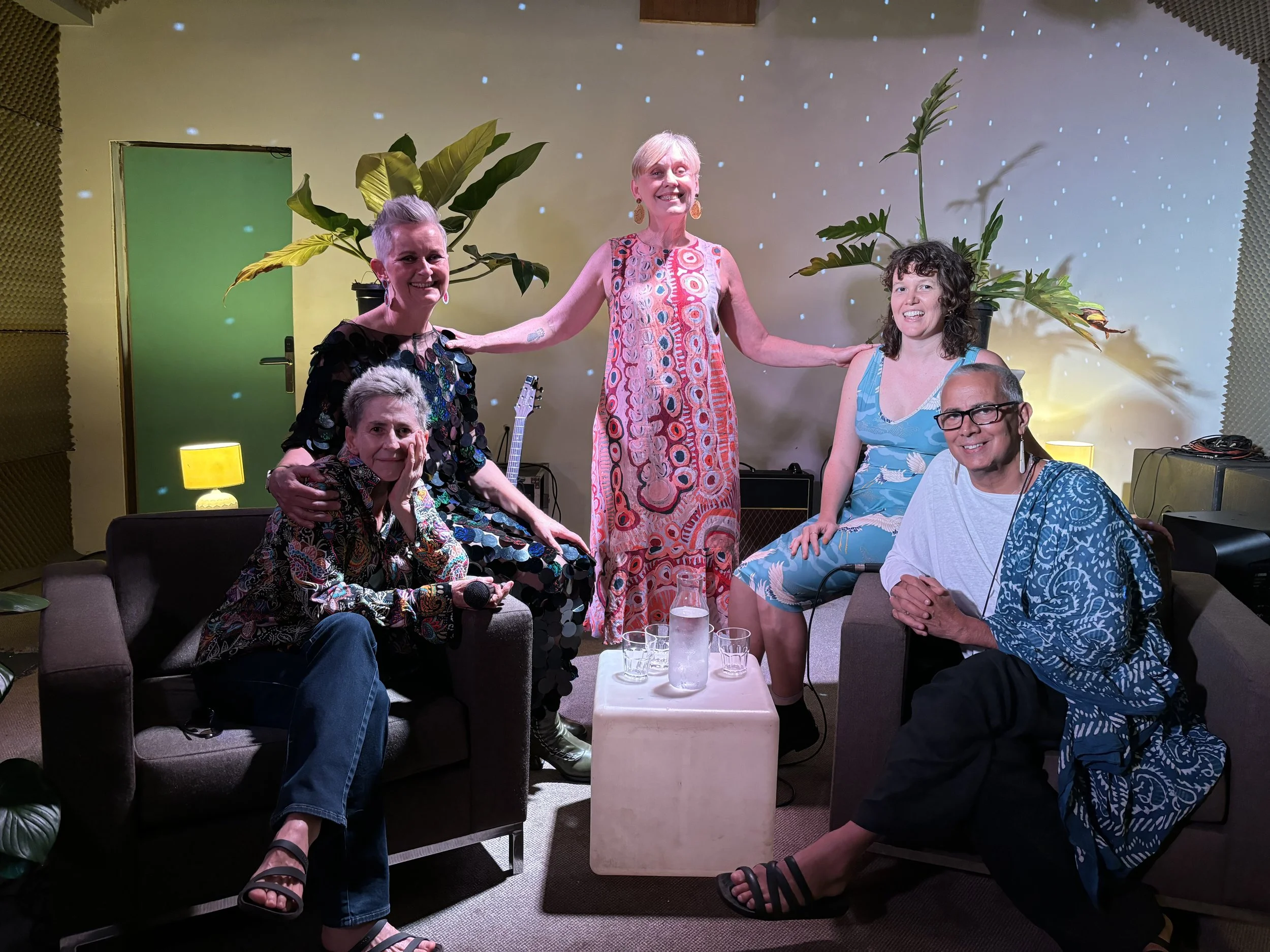 A group of five people, four women and one man, are gathered in a cozy indoor setting, sitting and standing around a small table with water glasses. The background features a wall with hanging plants, yellow lamps, and a starry projection. They are s
