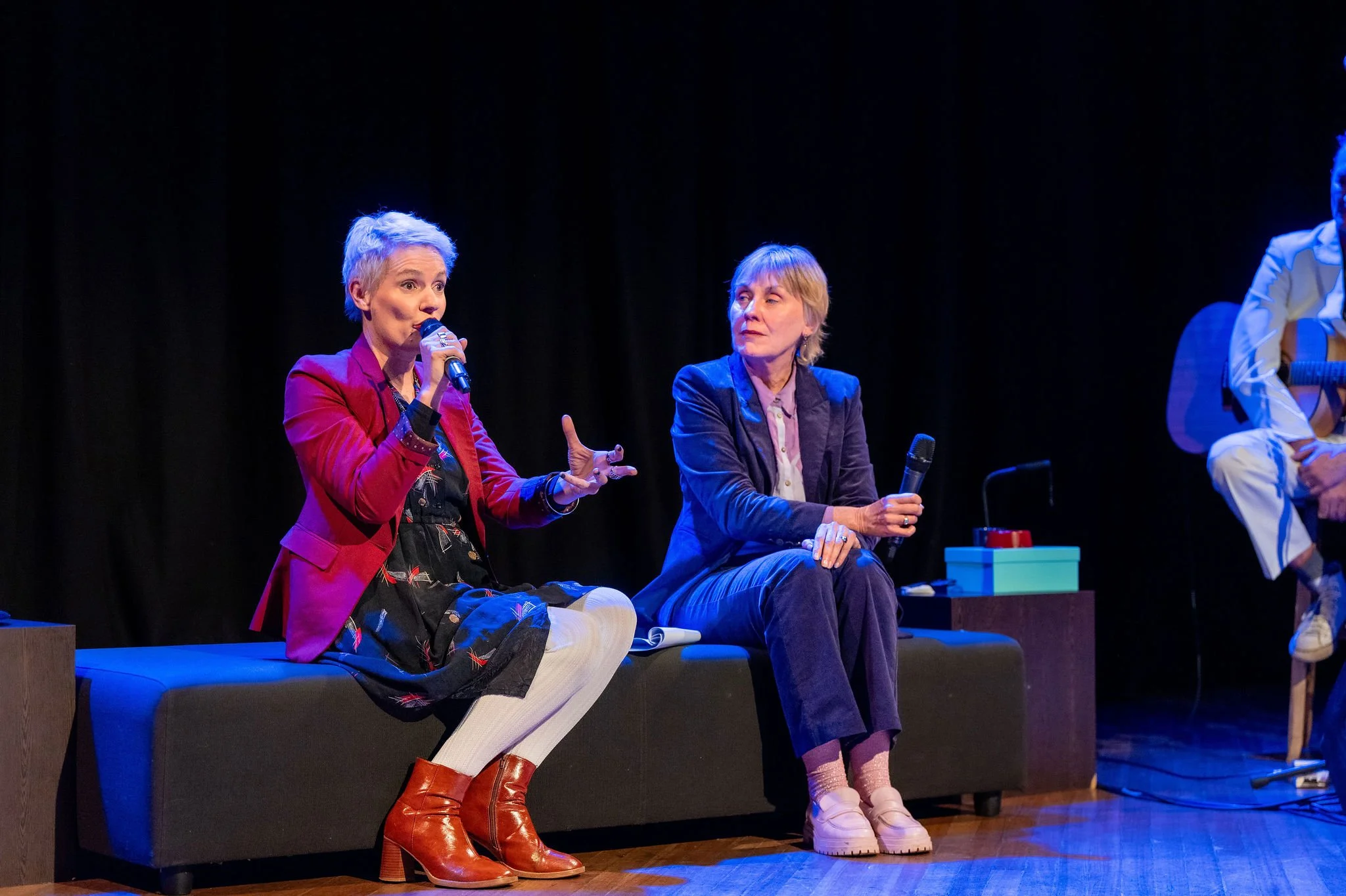 Two women seated on a stage, engaged in conversation during a panel discussion. One woman is speaking into a microphone while the other listens, holding a microphone in her lap. There is a person dressed as a clown in the background playing a guitar.