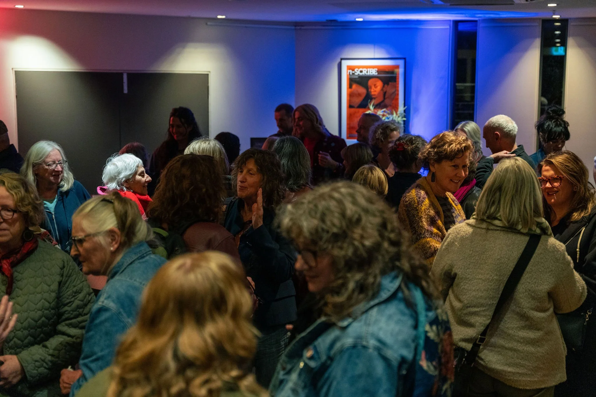 A crowded indoor social gathering with people smiling and talking, illuminated by colorful lights on the walls, with a framed artwork or poster hanging in the background.