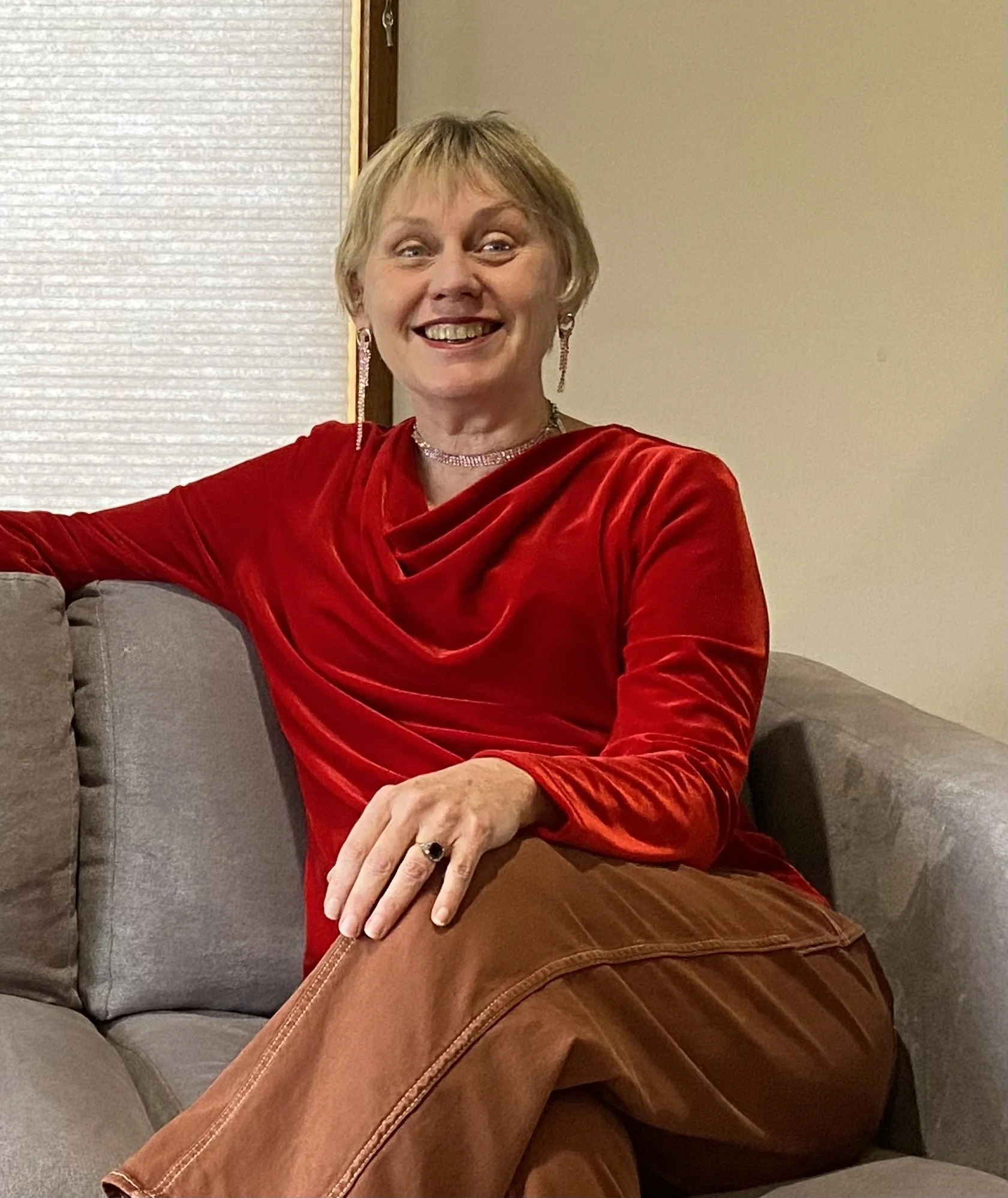 A woman with short blonde hair, wearing a red velvet top, chandelier earrings, a necklace, and a ring, smiling while sitting on a gray couch in a living room with beige walls and a window with blinds.