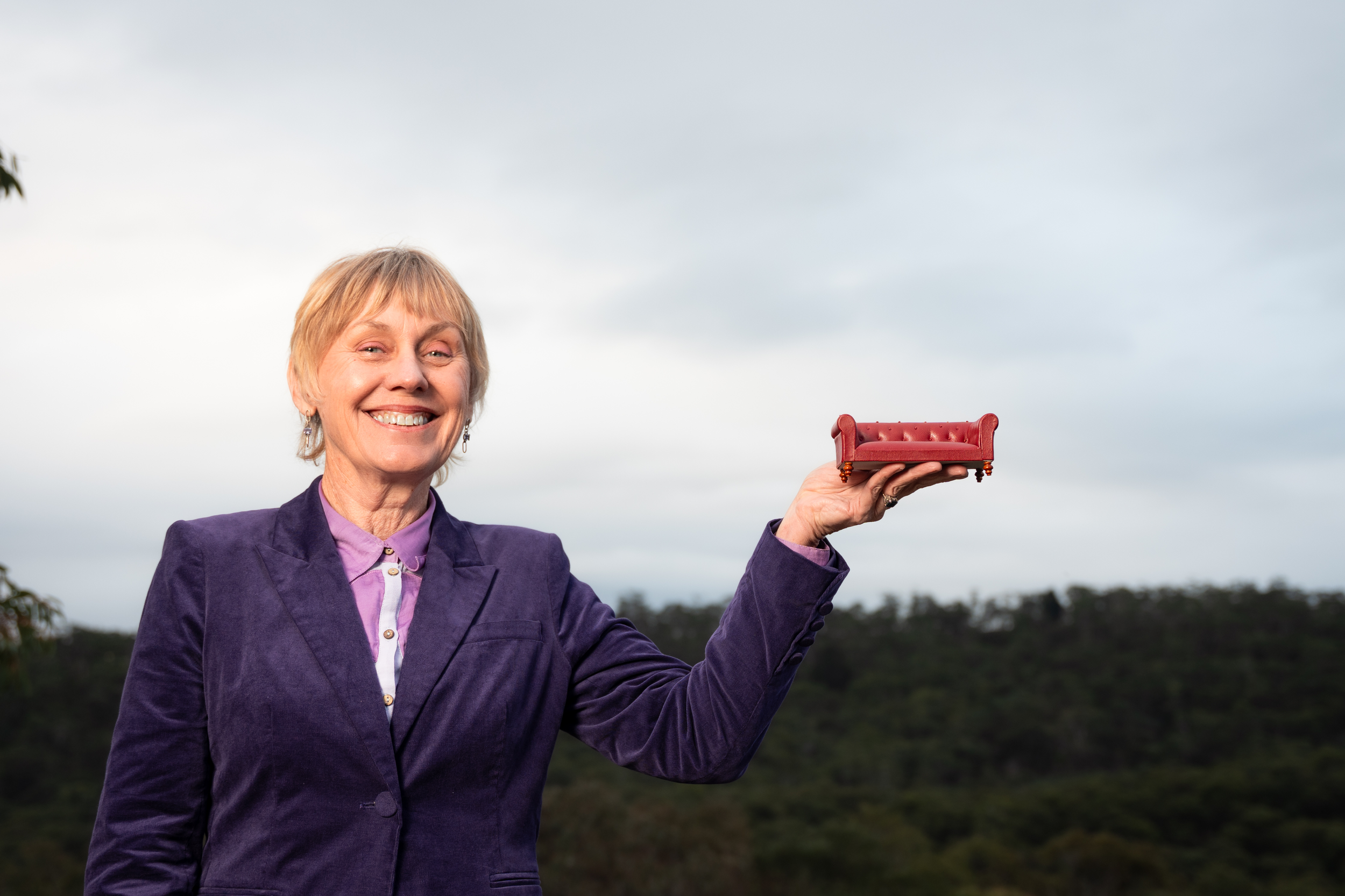 A smiling woman with short blonde hair holding a small red sofa figurine outdoors against a cloudy sky, dressed in a purple blazer and purple blouse.