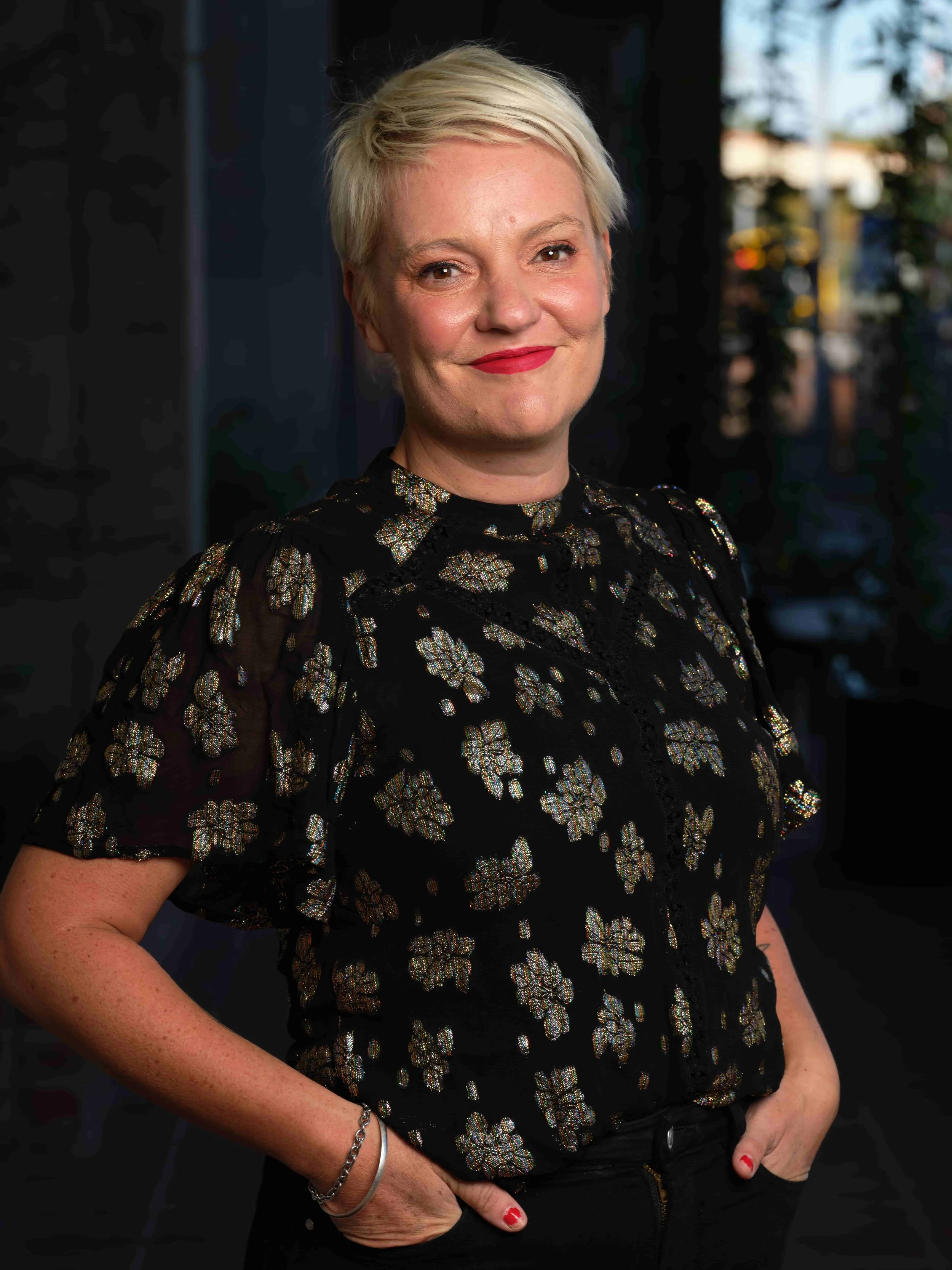 A woman with short platinum blonde hair, wearing a black top with metallic floral embroidery, standing indoors with a city street visible through a window behind her. She has red lipstick, a friendly expression, and is posing with hands in her pockets.