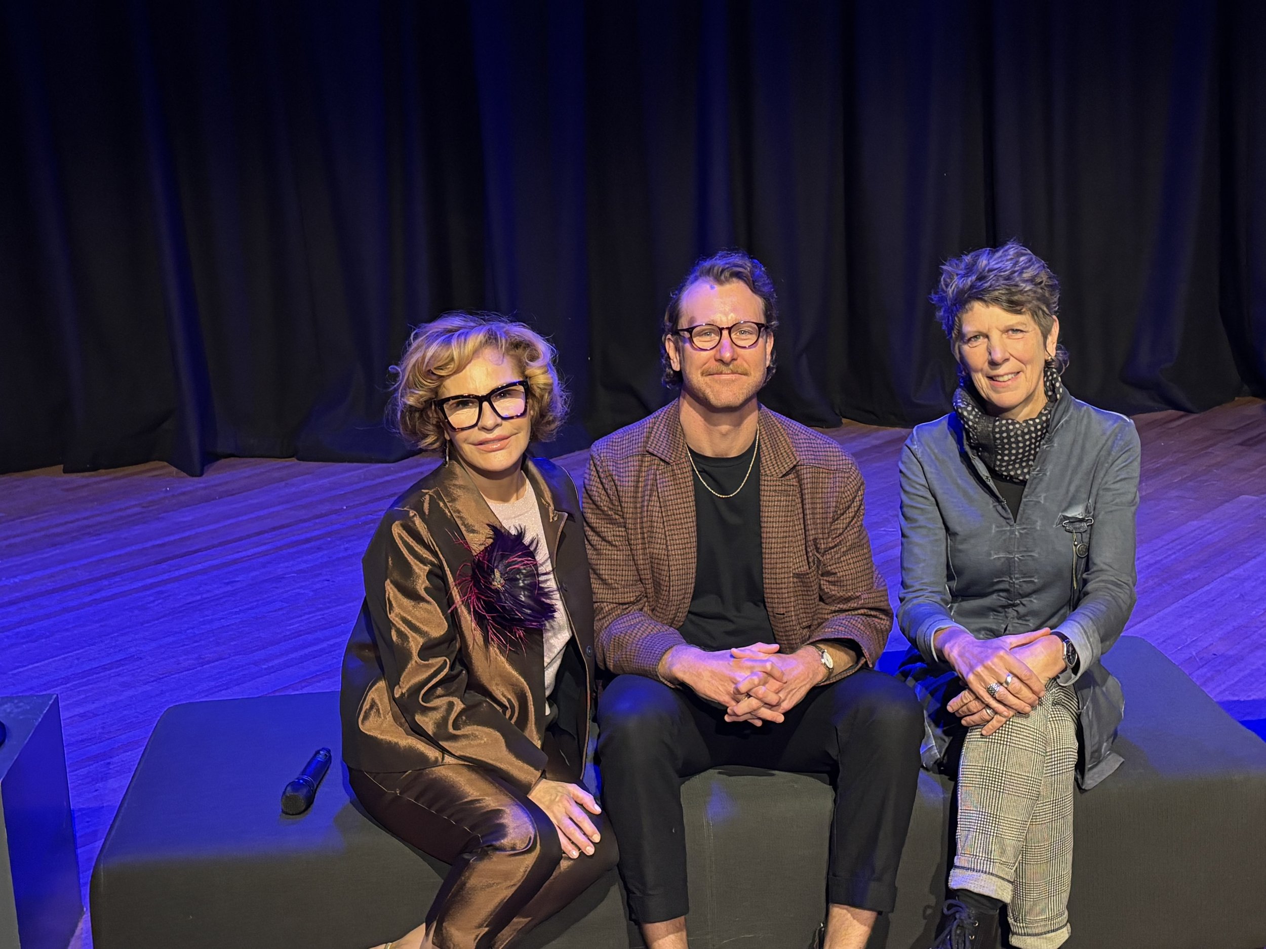 Three individuals sitting on chairs on a stage with a black curtain backdrop. The woman on the left has curly blonde hair and wears large glasses and a brown satin jacket. The man in the middle has dark hair, glasses, and wears a checkered blazer. Th