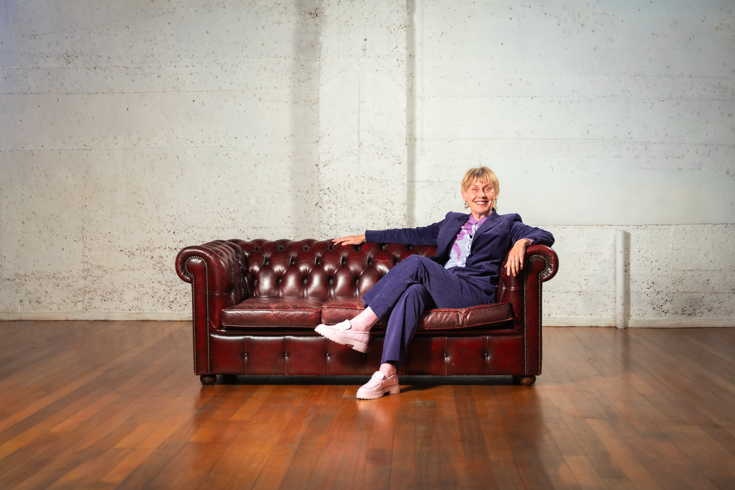 A woman with short blonde hair sitting on a red leather couch with wooden flooring and a plain white brick wall background. She is smiling, wearing a purple suit and pink sneakers, with one arm resting on the armrest.