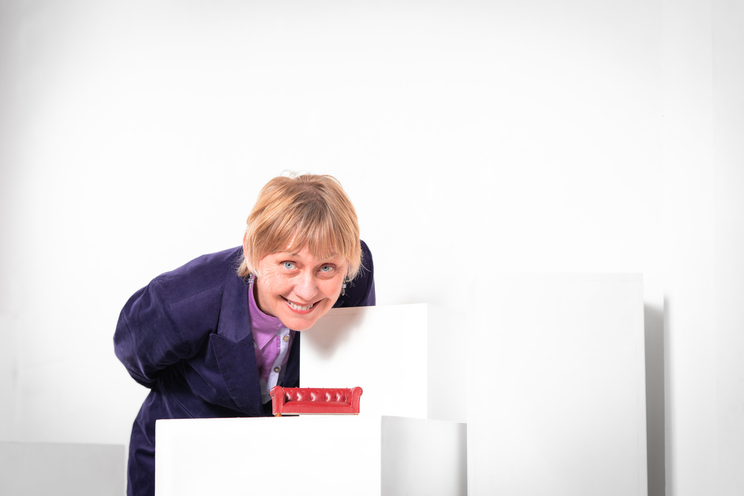A woman with short blonde hair and a big smile, wearing a purple shirt and dark blazer, leaning over a white table with a red stapler on it, against a plain white background.