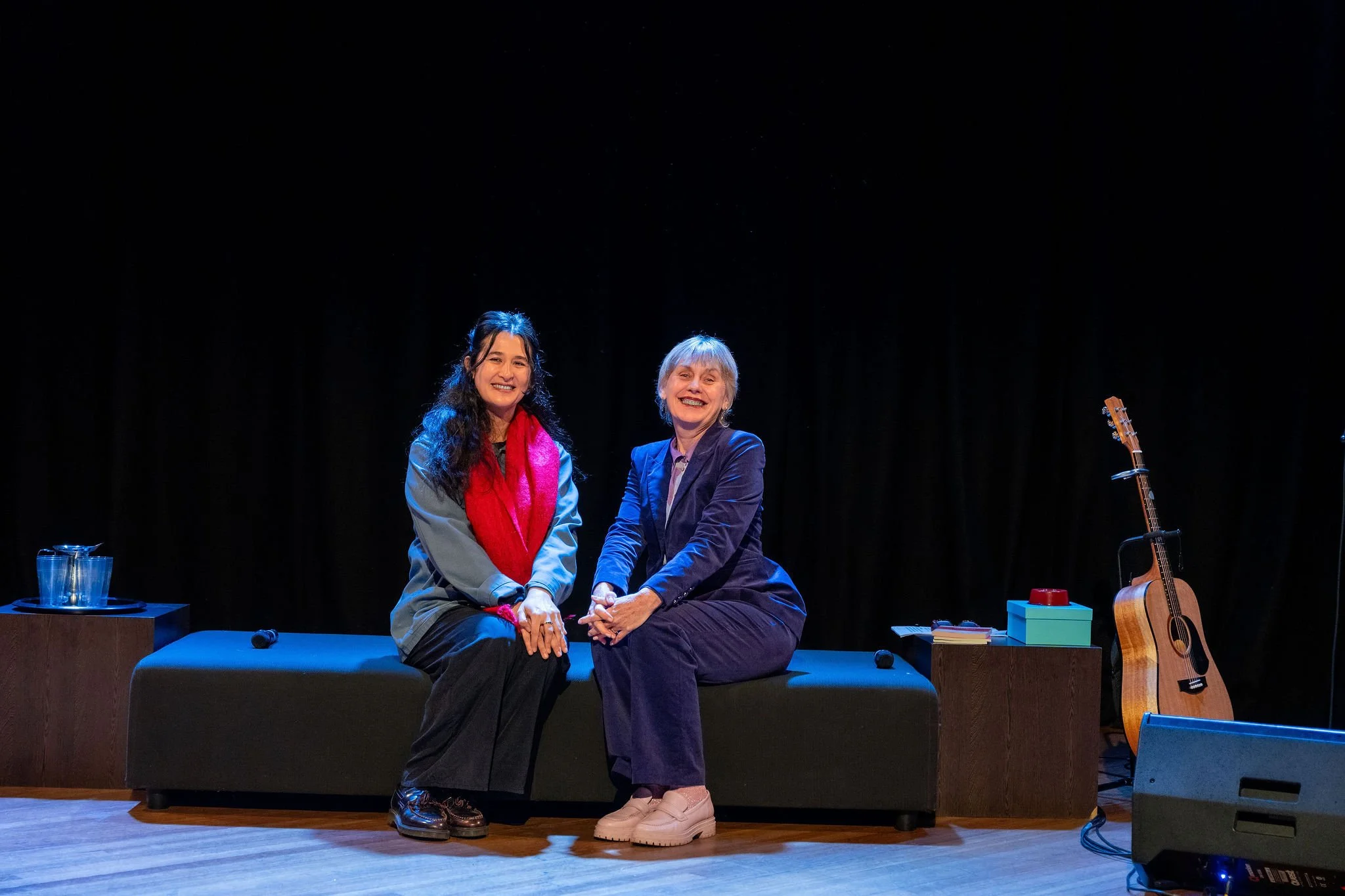 Two women sitting on a stage, holding hands and smiling, with a guitar and a small table with glasses nearby.