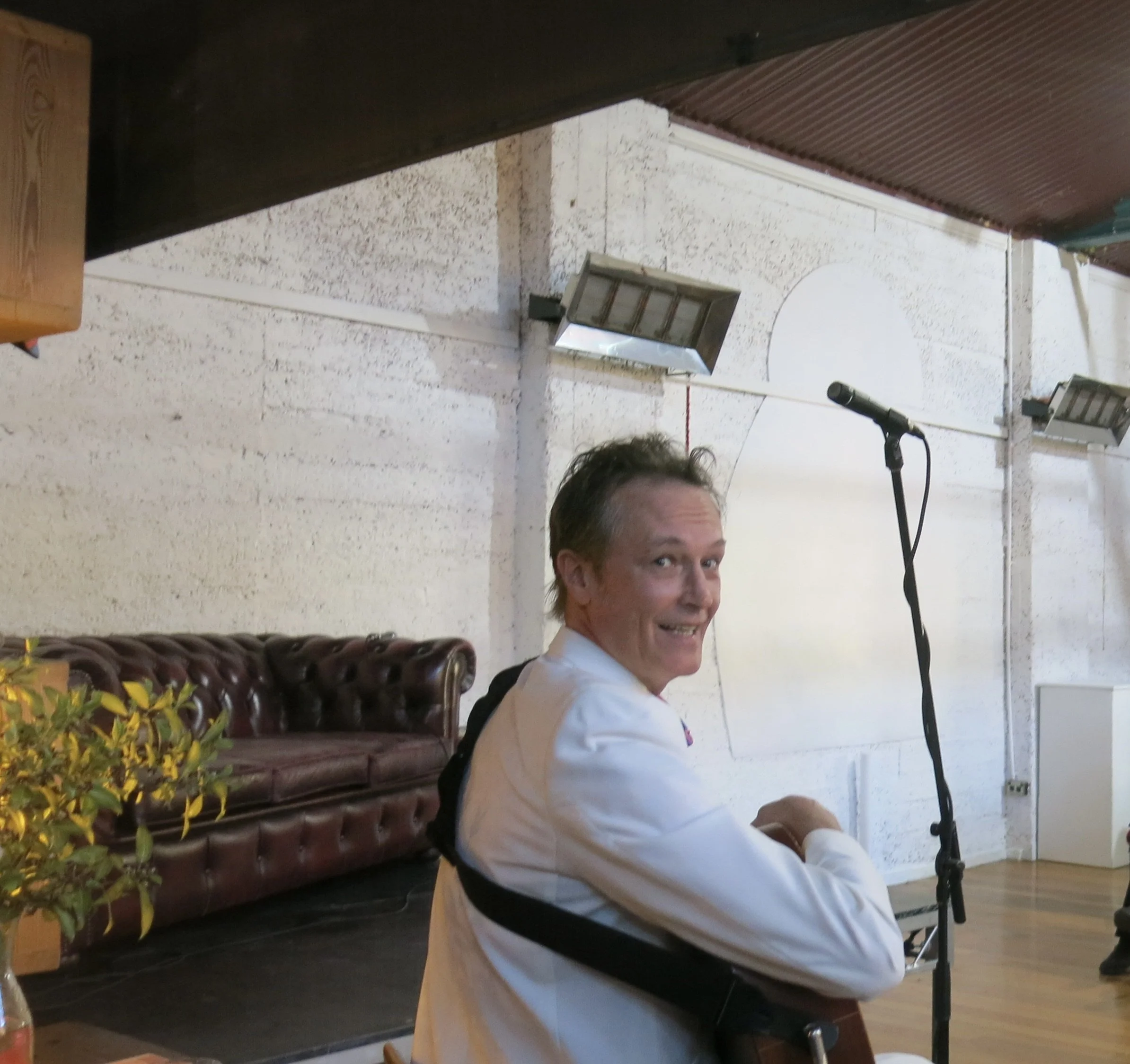 A man in a white shirt is smiling and looking at the camera, sitting in front of a microphone in an indoor setting with a textured white wall, two wall-mounted light fixtures, and a brown leather bench with yellow flowers in the foreground.