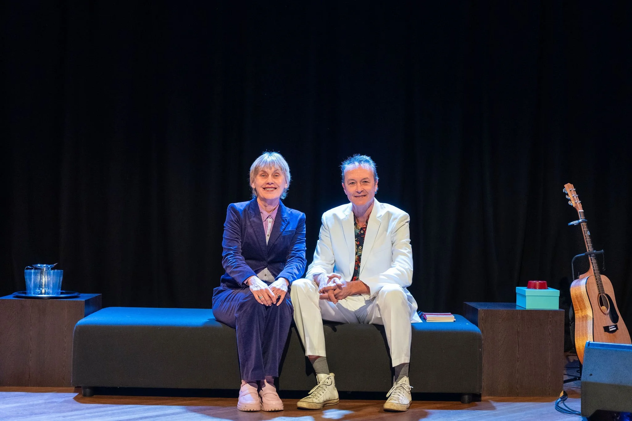 Two older men sitting on a stage with a black background. One wears a purple jacket, the other a white jacket. There are water glasses, a guitar, and colorful blocks on the stage.