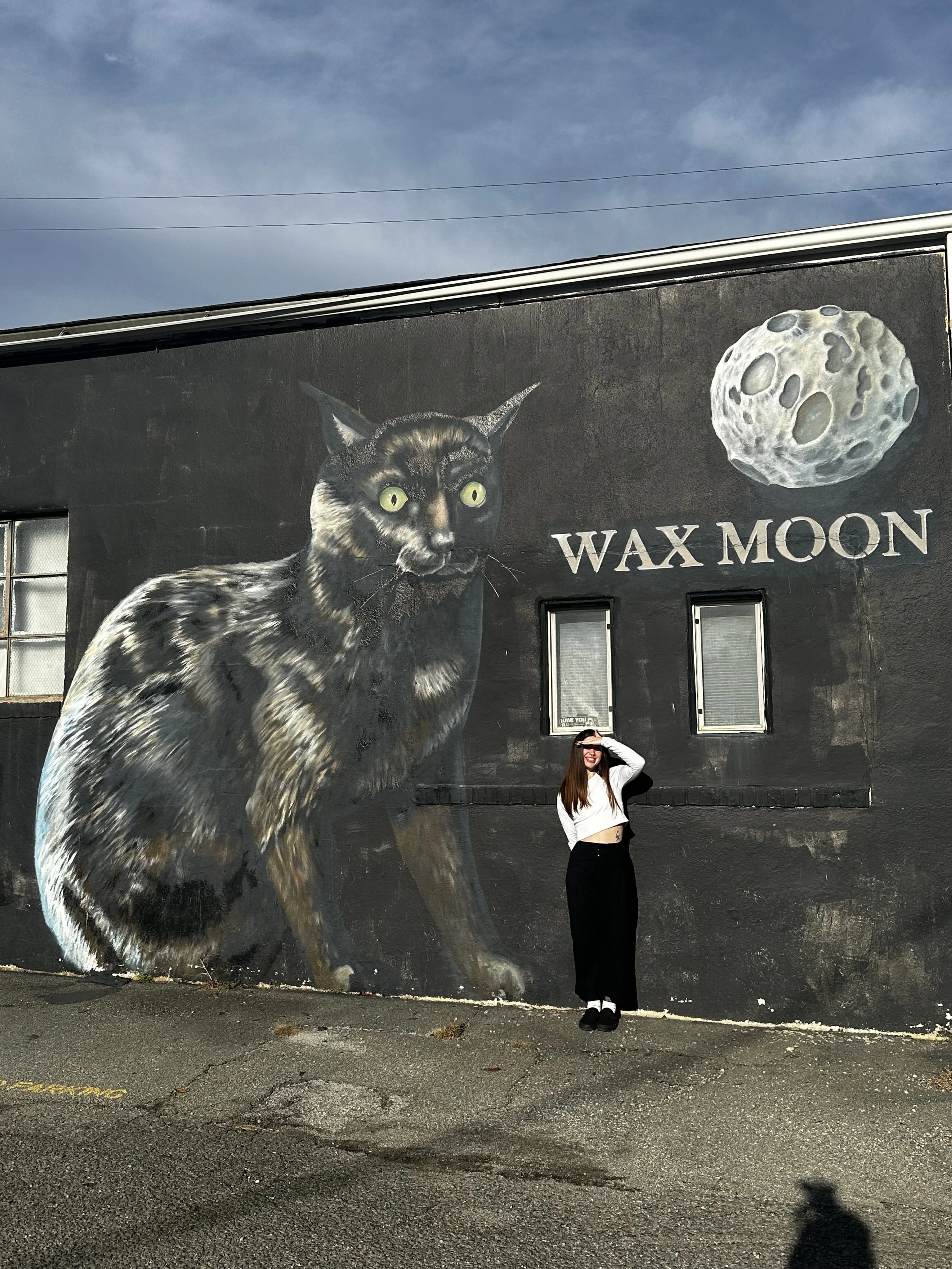 A mural of a black and brown cat with green eyes next to a depiction of the moon on an outdoor wall, with a young woman with long red hair, sunglasses, a white top, and black pants standing in front of it.